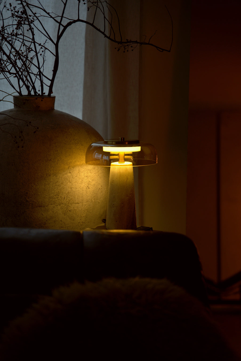 The Ankaa lamp lit beside a large stone vase, the rose glass glowing amber in a dark room.