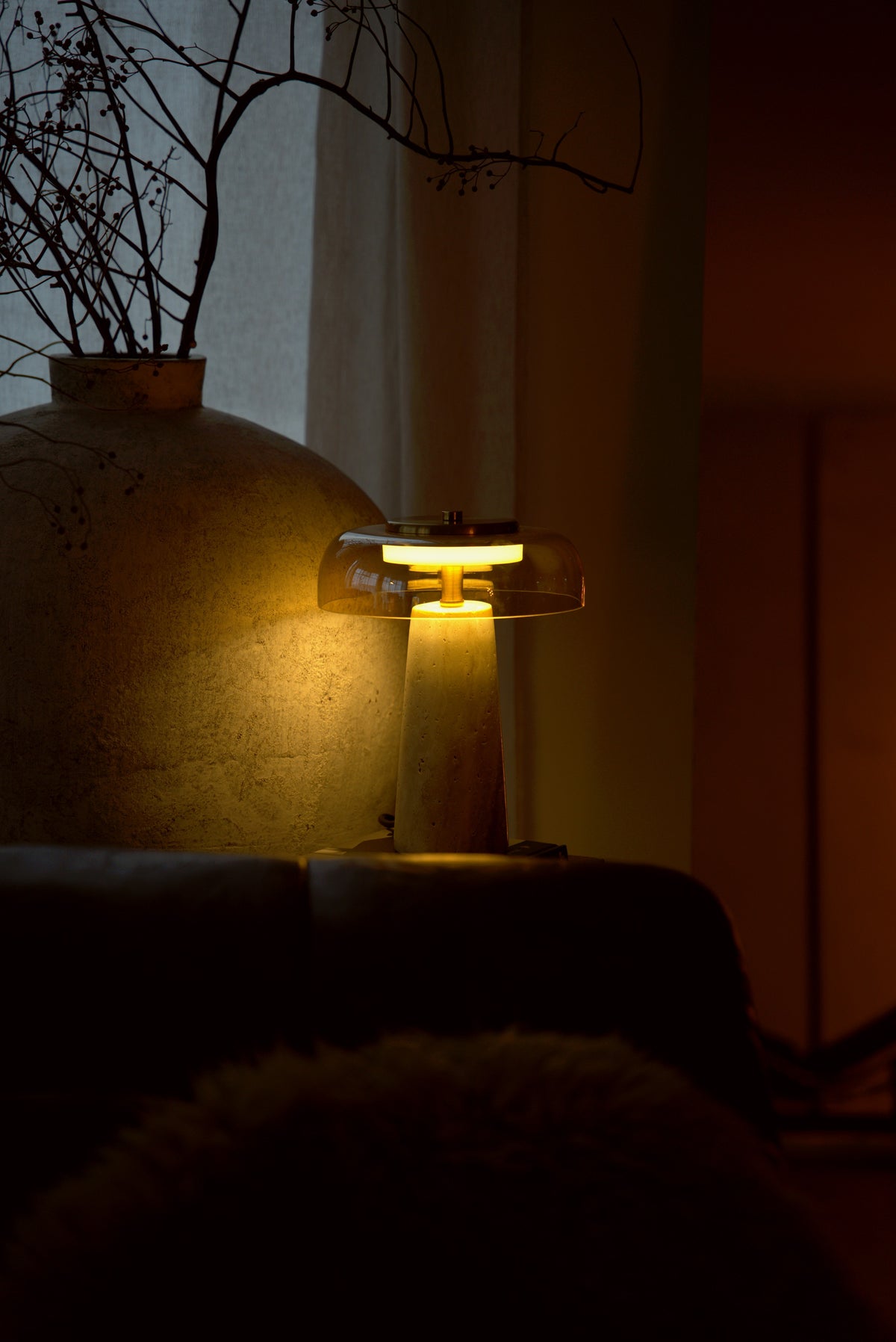 The Ankaa lamp lit beside a large stone vase, the rose glass glowing amber in a dark room.