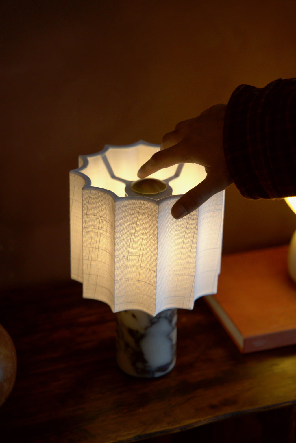 A hand at the Pavo lamp's brass finial, each pleat of the shade lit.