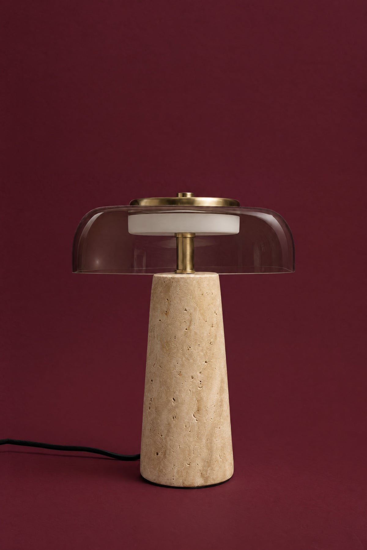 The Ankaa lamp, unlit: a wide, clear smoked glass shade over a warm honey travertine cone, brass stem and top plate, cord visible, against a deep oxblood background.