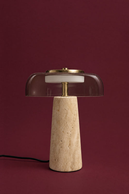 The Ankaa lamp, unlit: a wide, clear smoked glass shade over a warm honey travertine cone, brass stem and top plate, cord visible, against a deep oxblood background.