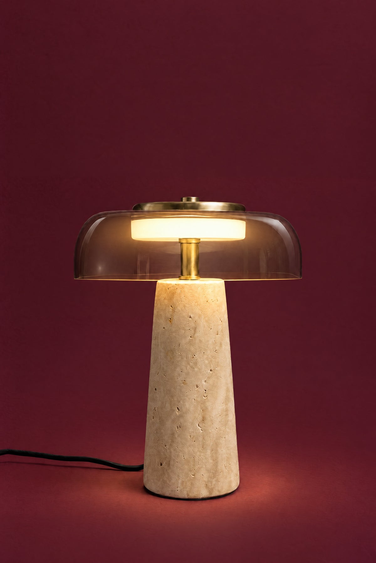 The Ankaa lamp, lit: the white disc inside the smoked glass glowing warm, light falling on the brass stem and the top of the travertine cone.