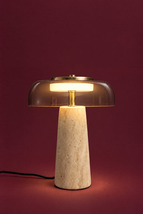 The Ankaa lamp, lit: the white disc inside the smoked glass glowing warm, light falling on the brass stem and the top of the travertine cone.