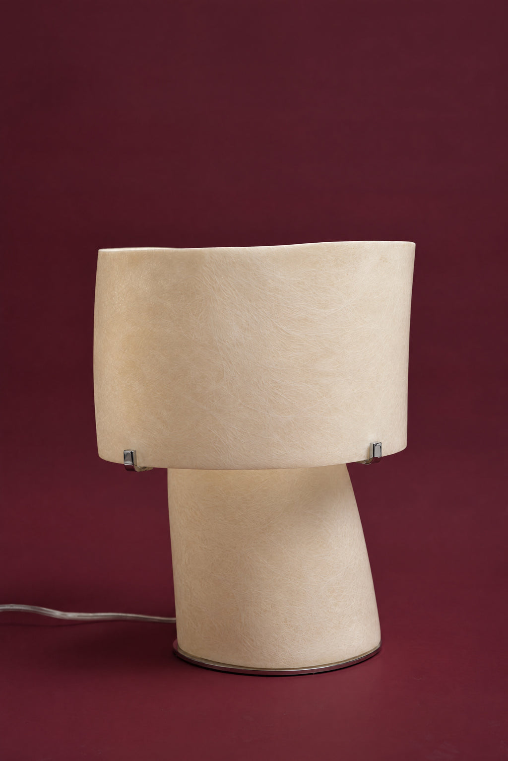 The Atria lamp, unlit: a cream fibreglass drum shade held by small chrome clips on a waisted fibreglass foot, its clear cord on the surface, against a deep oxblood background.