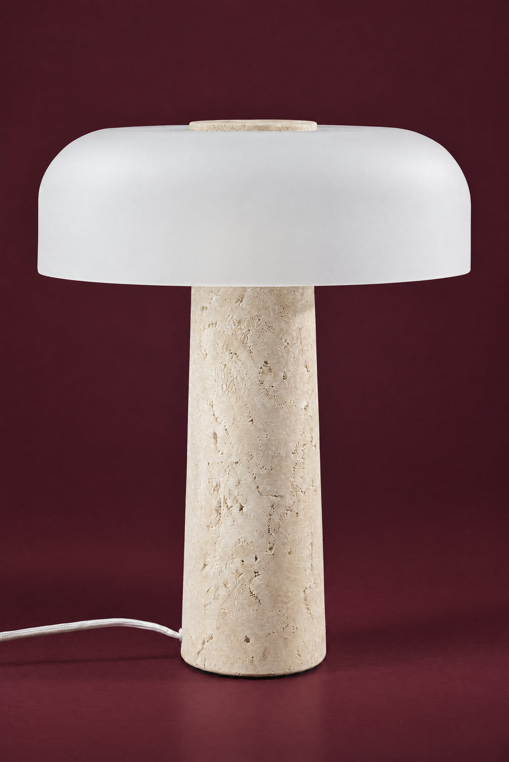 The Caelum lamp, unlit: a wide frosted white glass dome with a travertine disc in its crown, on a tall tapered travertine cone, against a deep oxblood background.