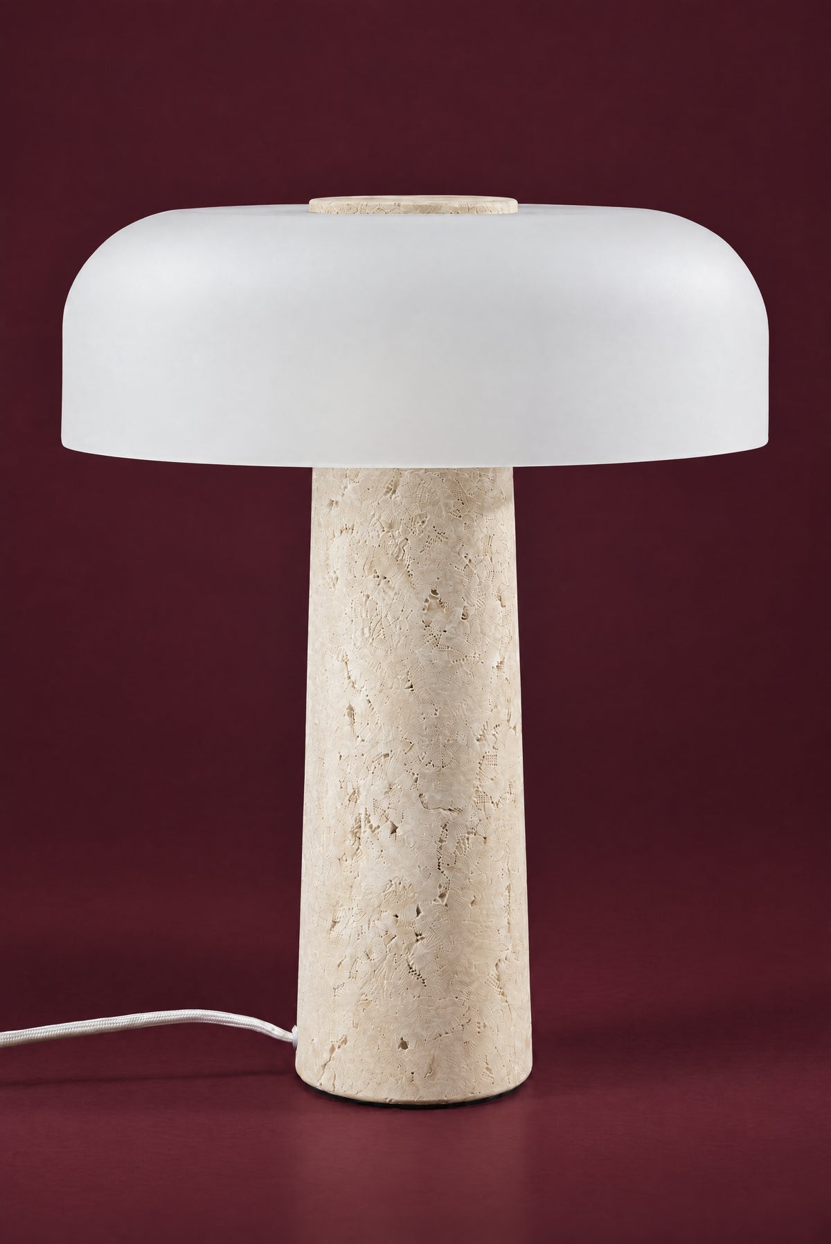 The Caelum lamp, unlit: a wide frosted white glass dome with a travertine disc in its crown, on a tall tapered travertine cone, against a deep oxblood background.