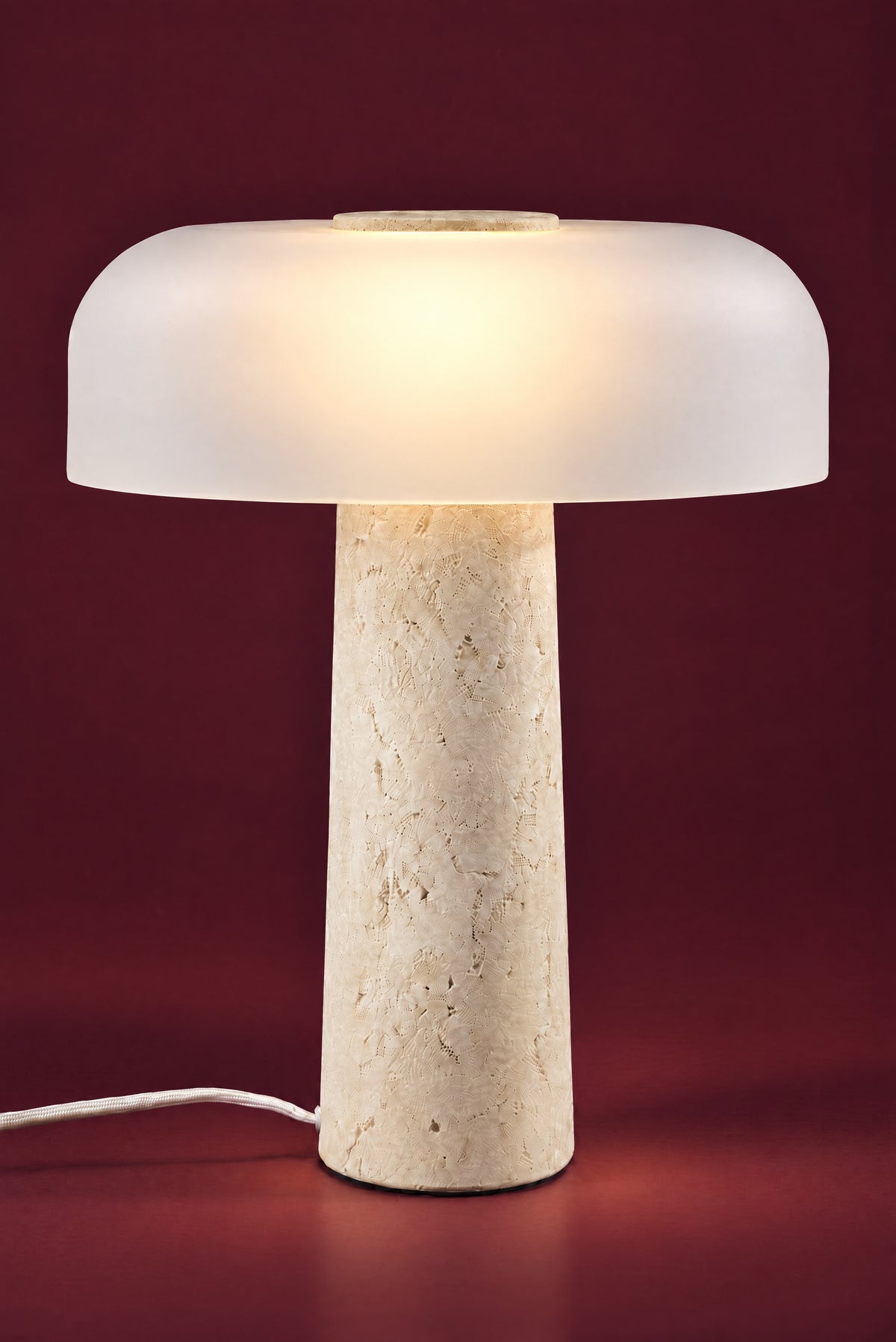 The Caelum lamp, lit: the frosted dome glowing warm from its centre, a soft pool of light on the floor around the travertine cone.