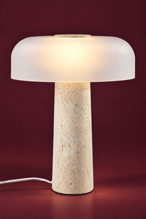 The Caelum lamp, lit: the frosted dome glowing warm from its centre, a soft pool of light on the floor around the travertine cone.