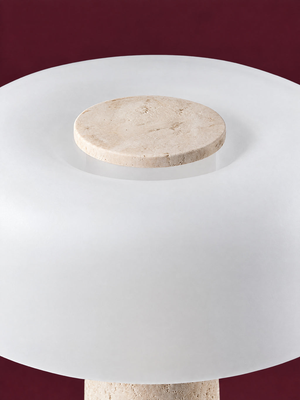 The Caelum lamp from above: the travertine disc set into the crown of the frosted glass dome, against a deep oxblood background.