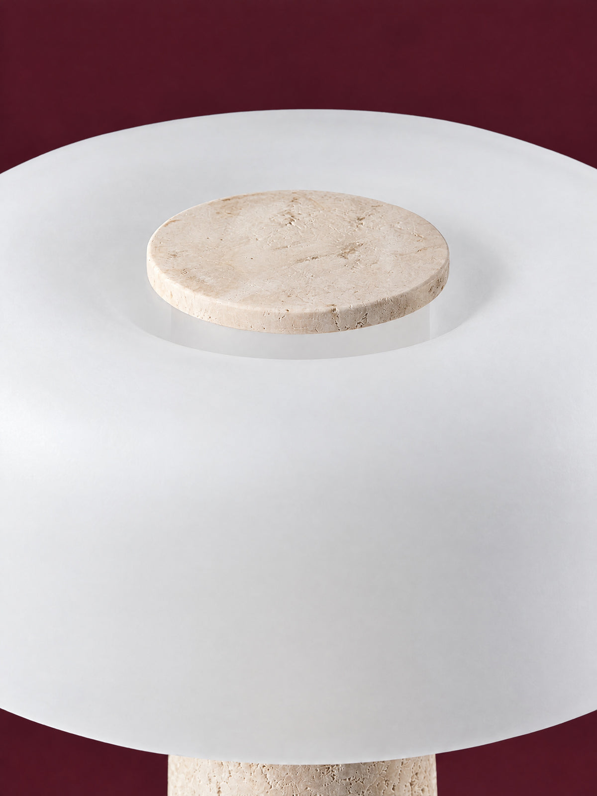 The Caelum lamp from above: the travertine disc set into the crown of the frosted glass dome, against a deep oxblood background.
