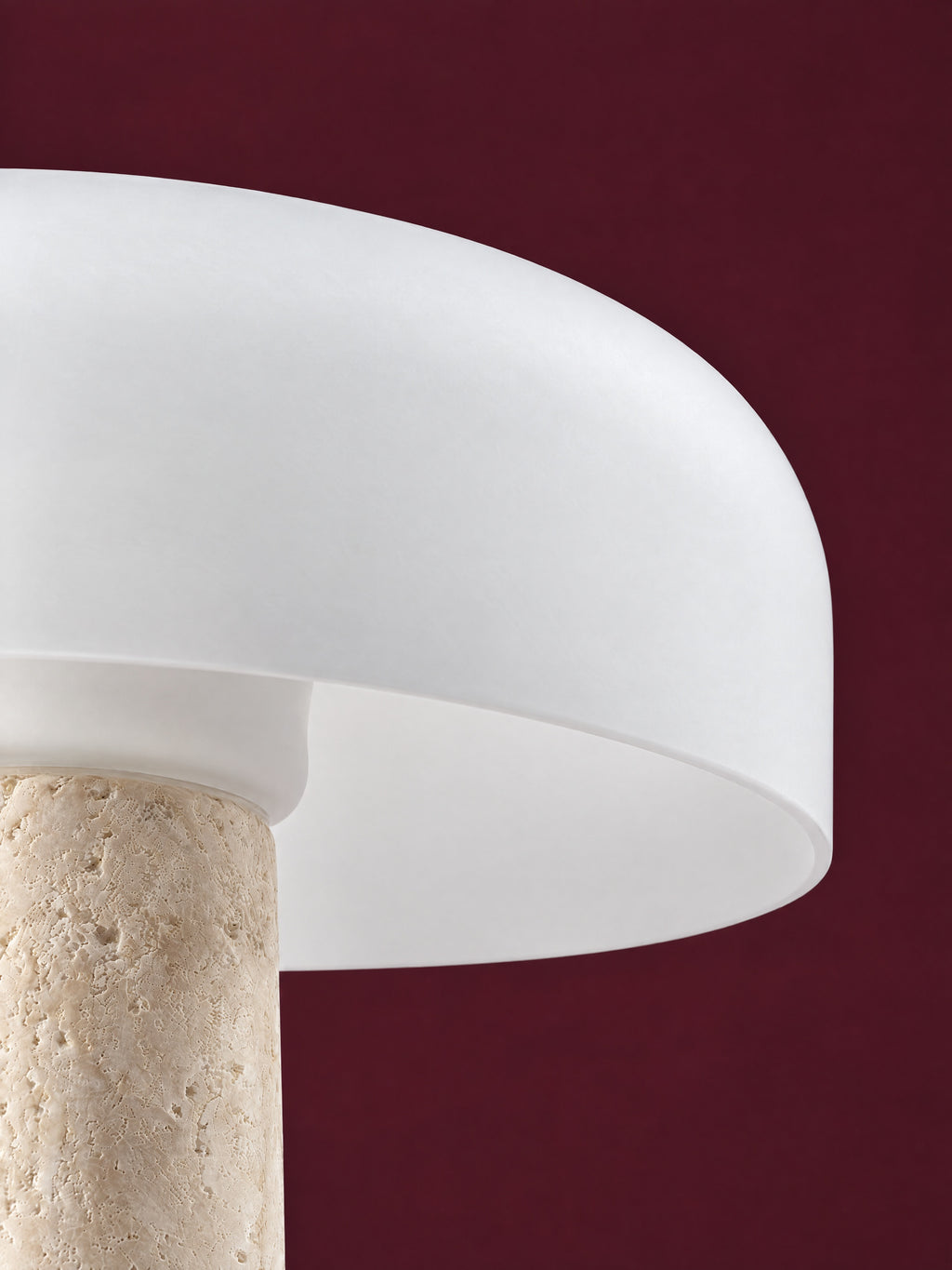 Detail of the Caelum lamp: the rim of the frosted glass dome where it meets the travertine cone, against a deep oxblood background.