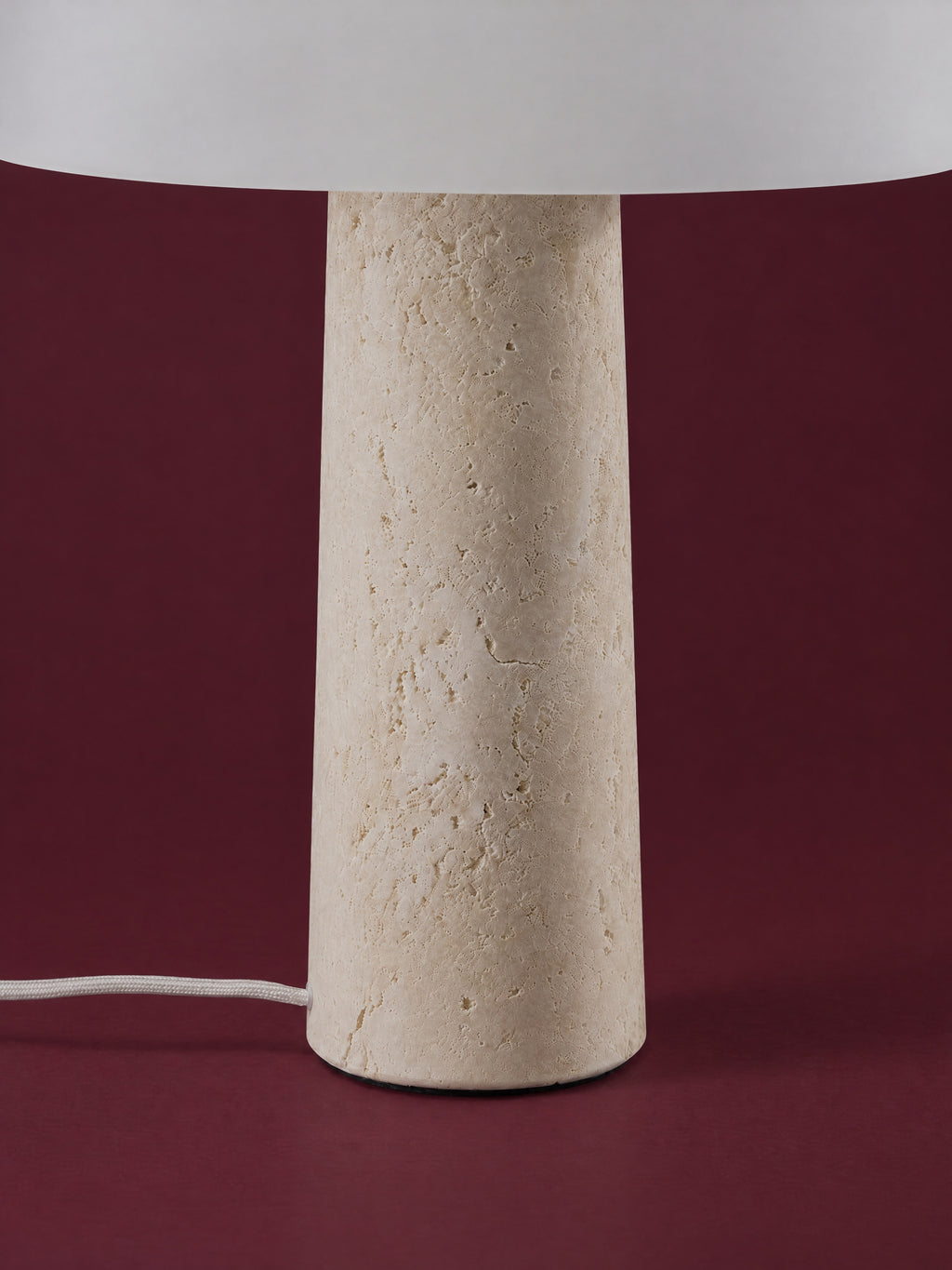 Detail of the Caelum lamp: the tapered travertine cone and its foot, the white cord on the floor, against a deep oxblood background.