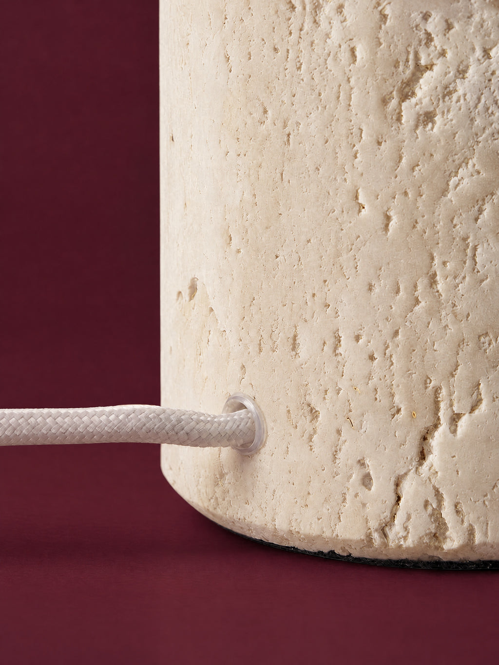 Detail of the Caelum lamp: the white braided cord entering the travertine cone just above its felt base, against a deep oxblood background.