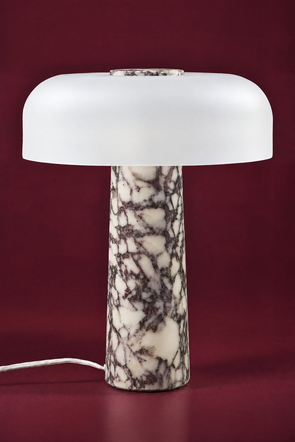 The Caelum lamp in Calacatta Viola, unlit: a wide frosted white glass dome with a marble disc in its crown, on a tall tapered cream and plum-veined marble cone, against a deep oxblood background.