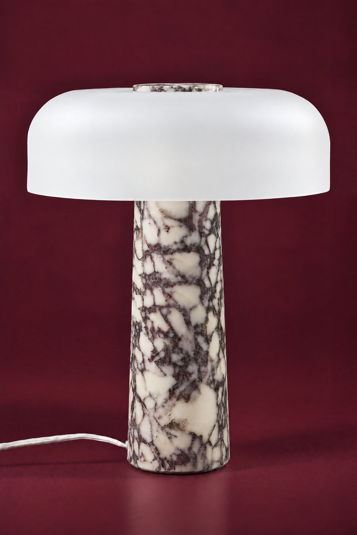 The Caelum lamp in Calacatta Viola, unlit: a wide frosted white glass dome with a marble disc in its crown, on a tall tapered cream and plum-veined marble cone, against a deep oxblood background.