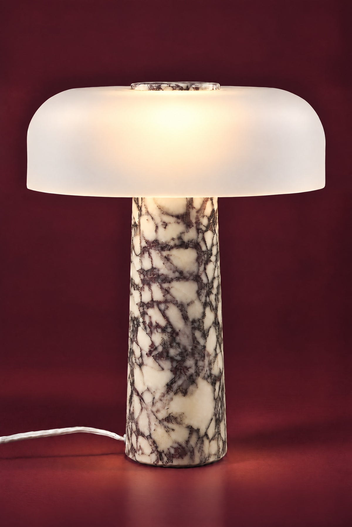 The Caelum lamp in Calacatta Viola, lit: the frosted dome glowing warm from its centre, a soft pool of light on the floor around the veined marble cone.