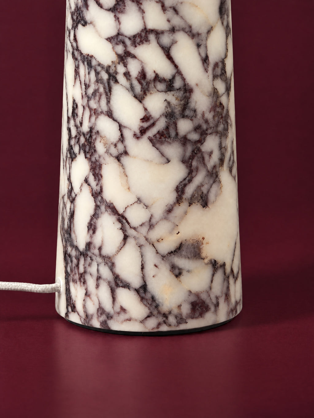Detail of the Caelum lamp in Calacatta Viola: the polished marble cone and its foot, the white braided cord entering just above the base, against a deep oxblood background.