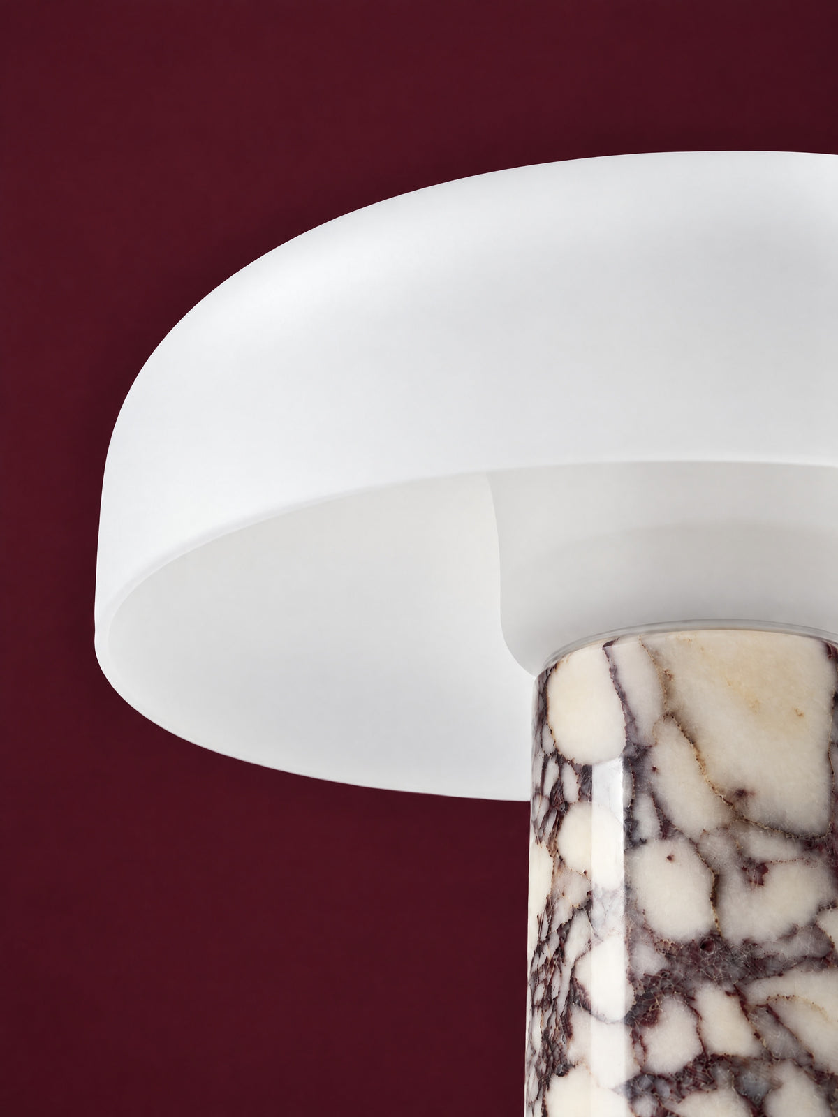 Detail of the Caelum lamp in Calacatta Viola: the rim of the frosted glass dome where it meets the marble cone, against a deep oxblood background.