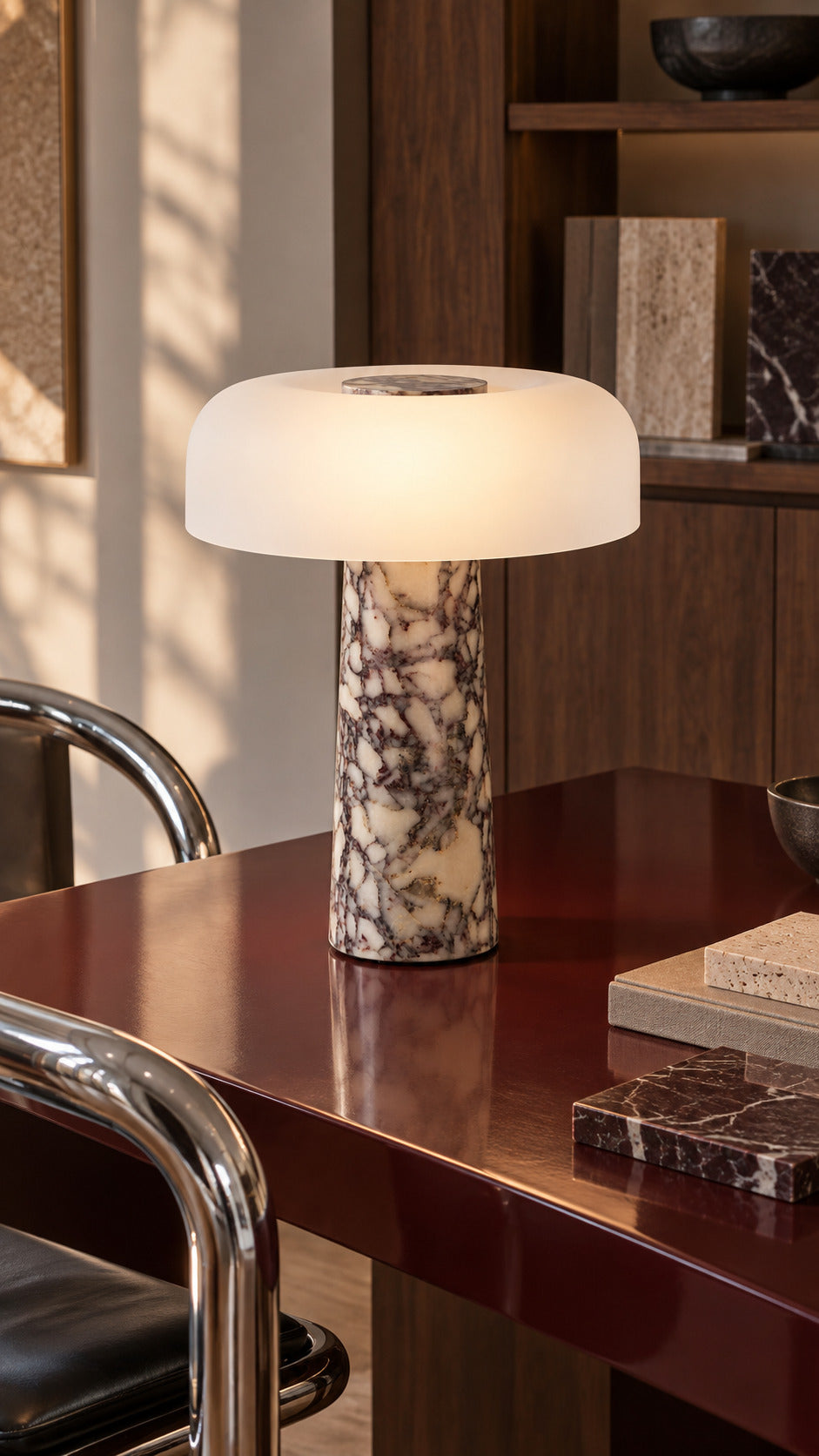 The Caelum lamp in Calacatta Viola, lit on an oxblood lacquered worktable in a material library, walnut sample drawers and a chrome chair behind