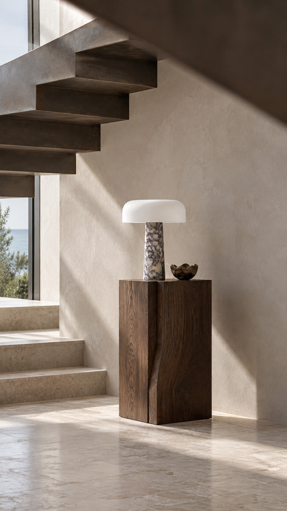 The Caelum lamp in Calacatta Viola, unlit on a walnut pedestal in a coastal stair hall, a folded-steel stair overhead and lime-plaster walls
