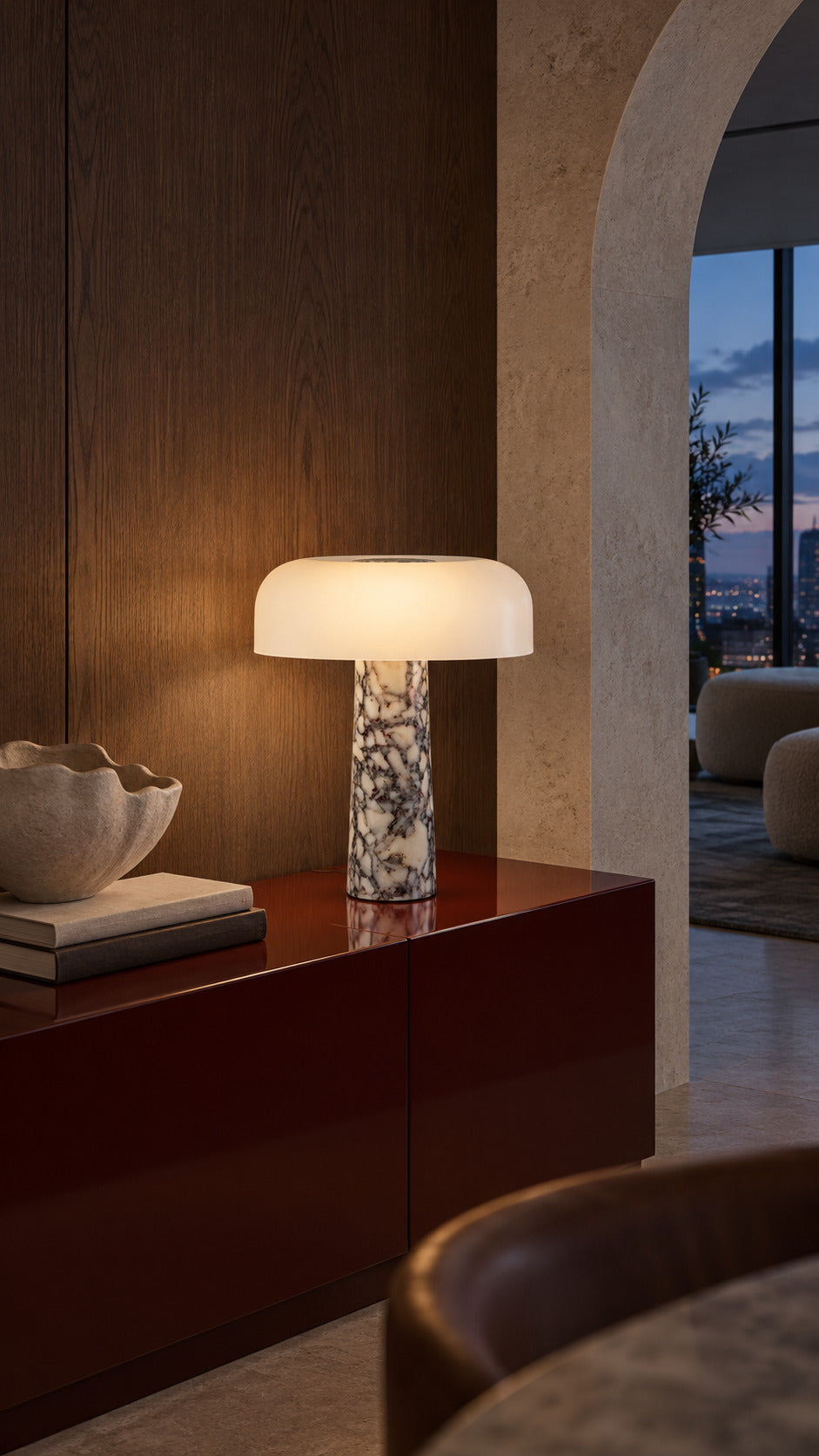 The Caelum lamp in Calacatta Viola, lit on an oxblood lacquer sideboard at blue hour, smoked oak panelling and an arched opening to the city lights