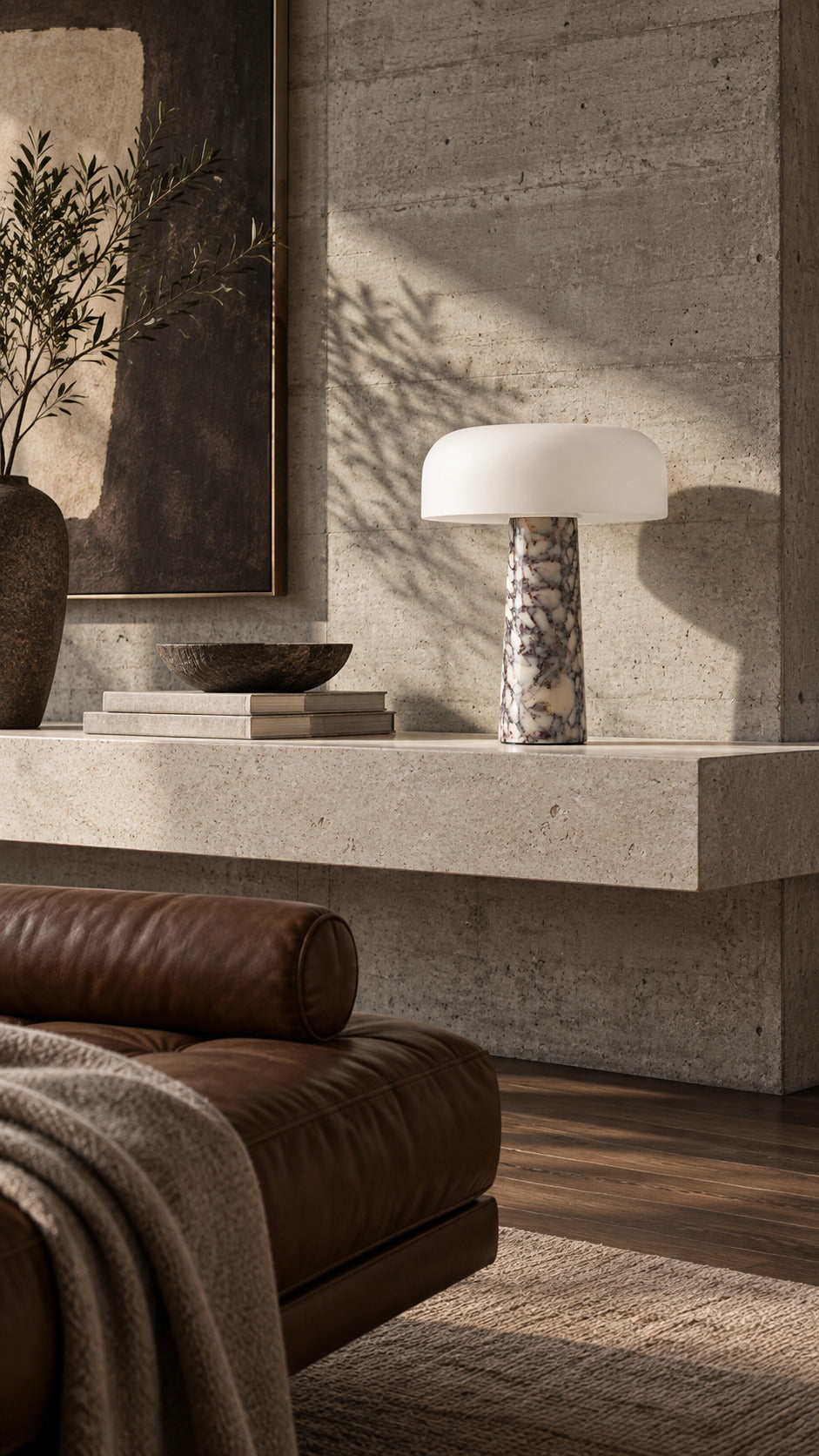 The Caelum lamp in Calacatta Viola, unlit on a floating limestone shelf against board-formed concrete, a tobacco leather daybed below