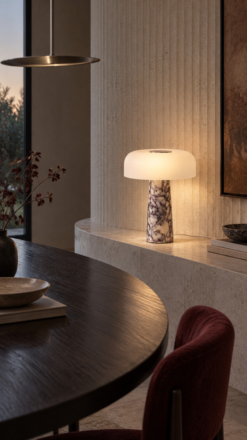 The Caelum lamp in Calacatta Viola, lit on a limestone ledge against a fluted stone wall, seen across a dark elliptical dining table at dusk