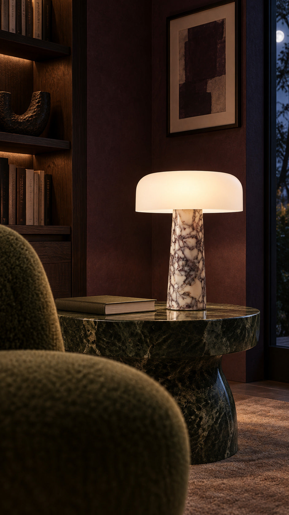 The Caelum lamp in Calacatta Viola, lit at night on a dark green serpentine side table, a smoked oak library and moss mohair chairs