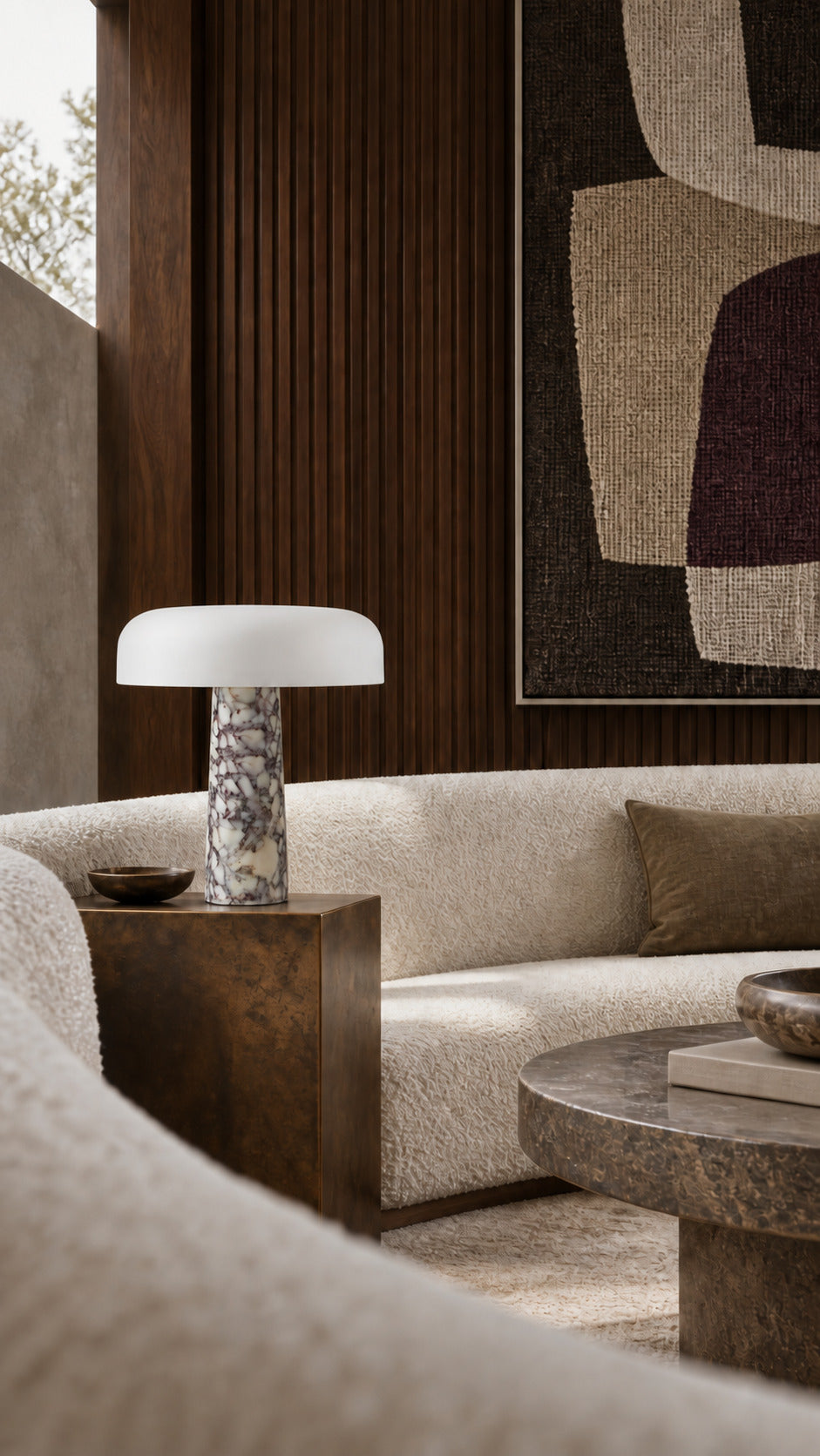 The Caelum lamp in Calacatta Viola, unlit on a patinated bronze plinth beside a curved boucle sofa, walnut ribs and a woven artwork behind