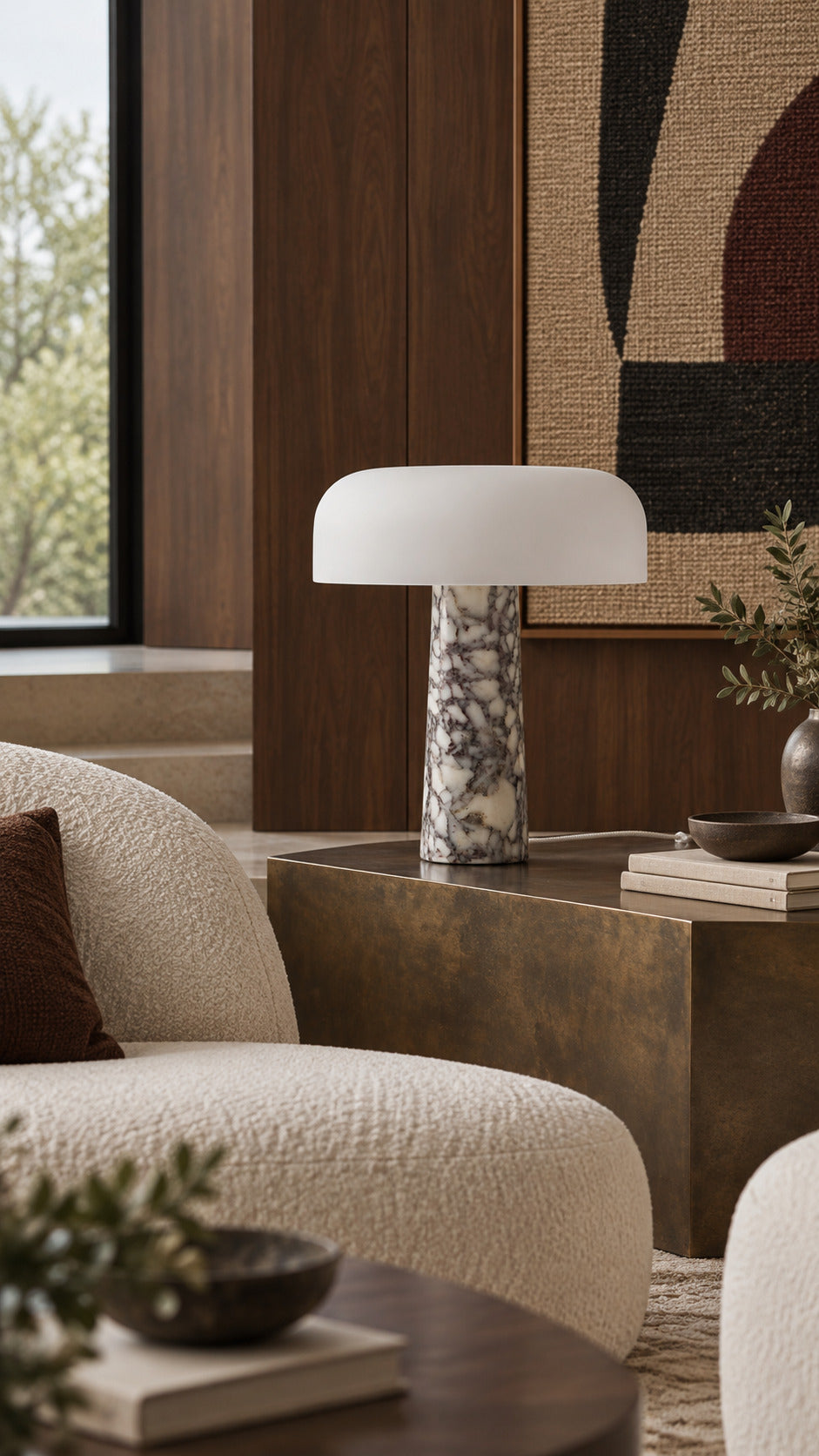 The Caelum lamp in Calacatta Viola, unlit on a patinated bronze cube between boucle armchairs, a woven artwork and a garden window behind