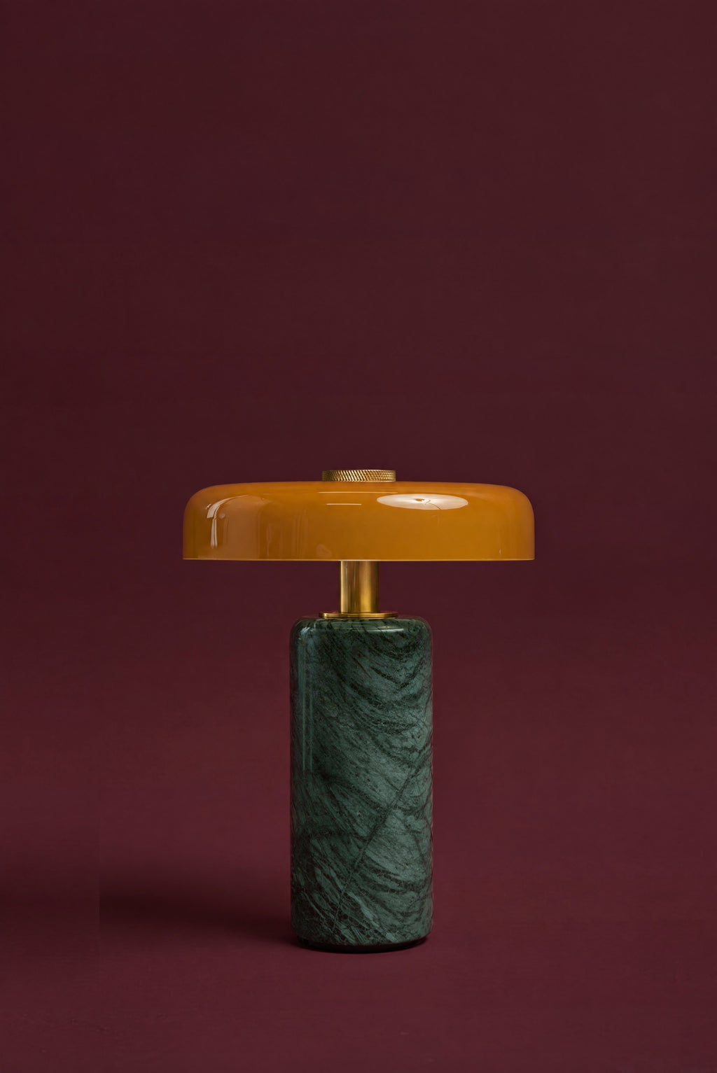 The Columba lamp in amber glass, unlit: an amber glass disc on a brass stem above a dark green Verde marble cylinder, against a deep oxblood background.