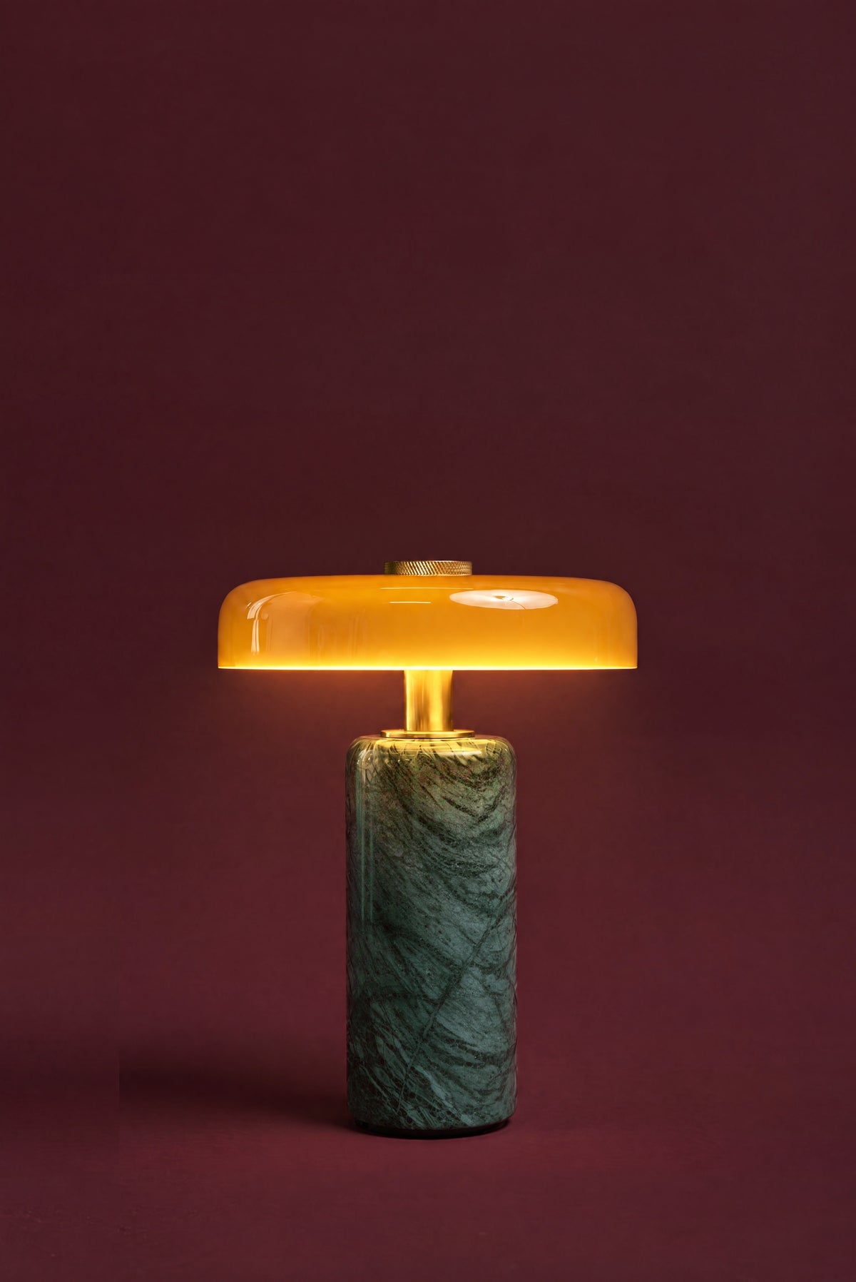The Columba lamp in amber glass, lit: the amber disc glowing honey-gold, warm light falling on the brass stem and the green marble column.