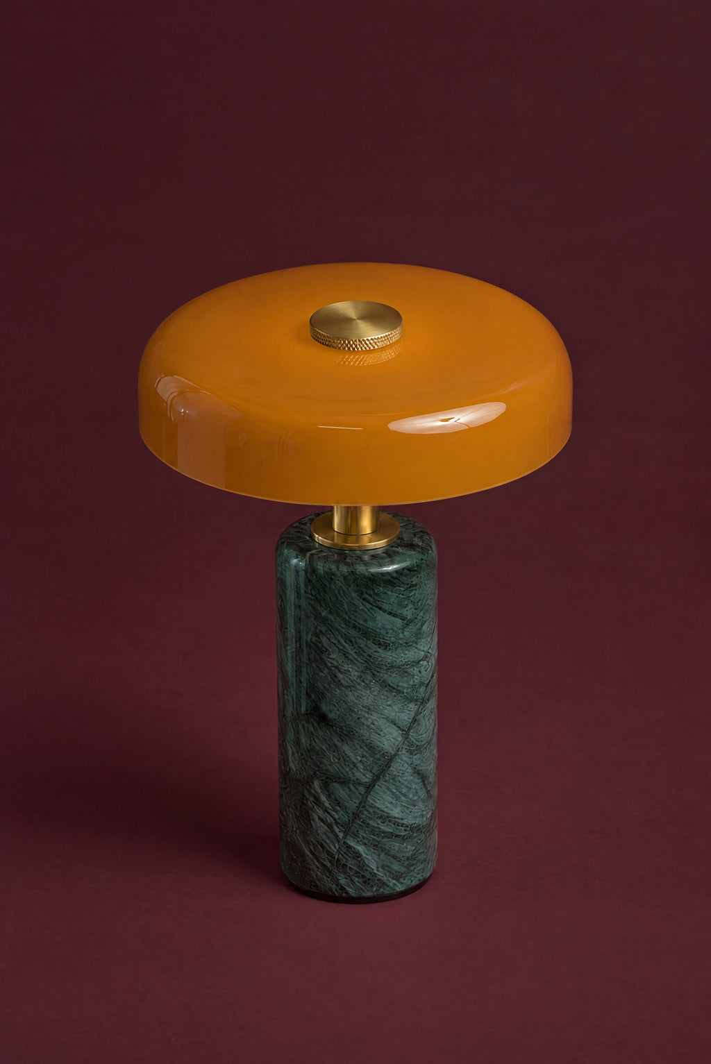 The Columba lamp in amber glass and Verde marble from above: the glossy disc with its knurled brass touch cap, over the dark green marble column, against a deep oxblood background.