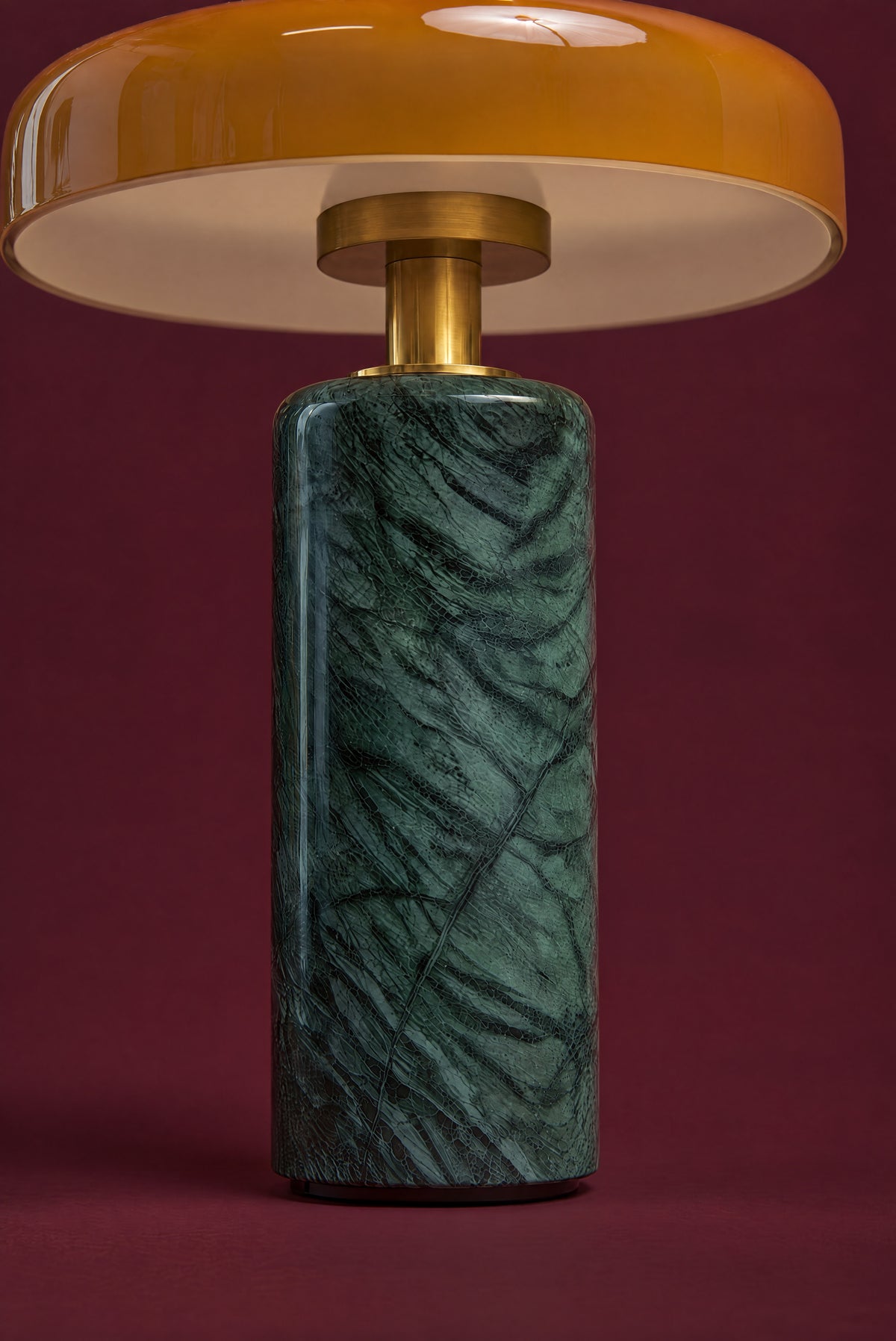 Under the disc of the Columba lamp, seen straight on: the cream-white inside of the amber glass shade, the brass cup and stem, and the brass collar on the green marble column.