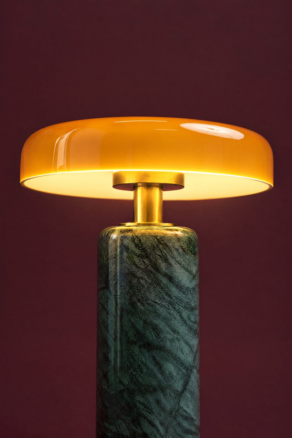 The Columba lamp lit: the amber glass disc glowing warm, light falling on the brass stem and the top of the green marble column.