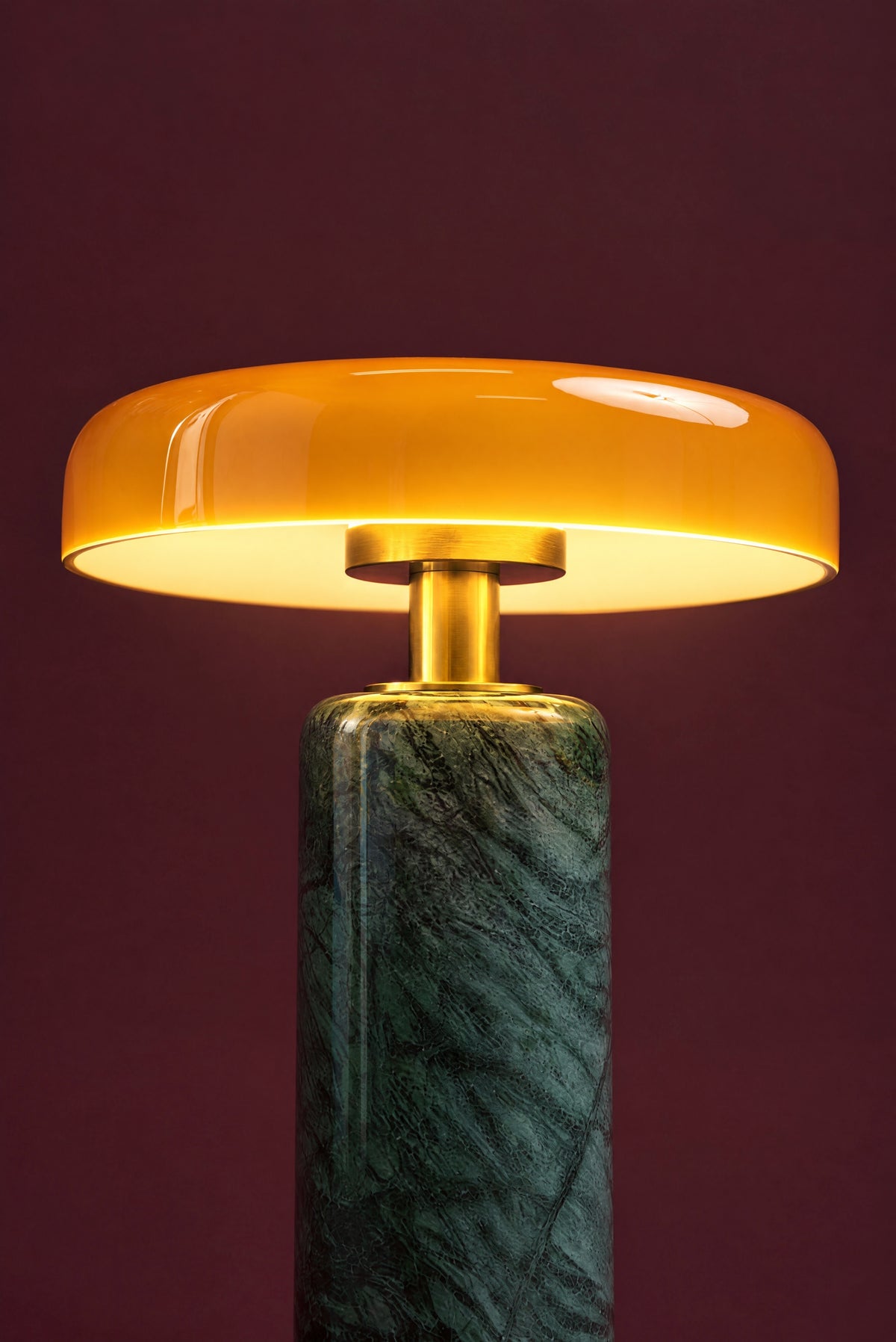 The Columba lamp lit: the amber glass disc glowing warm, light falling on the brass stem and the top of the green marble column.