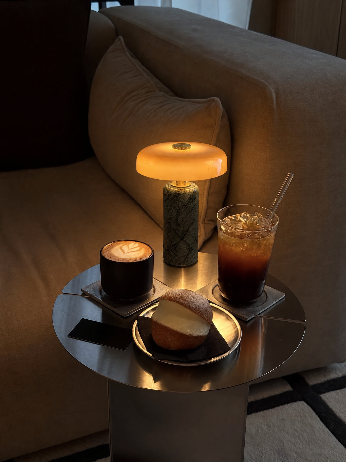 The Columba lamp lit on a round steel side table beside a linen sofa, with a latte, an iced coffee and a cream bun in the evening.