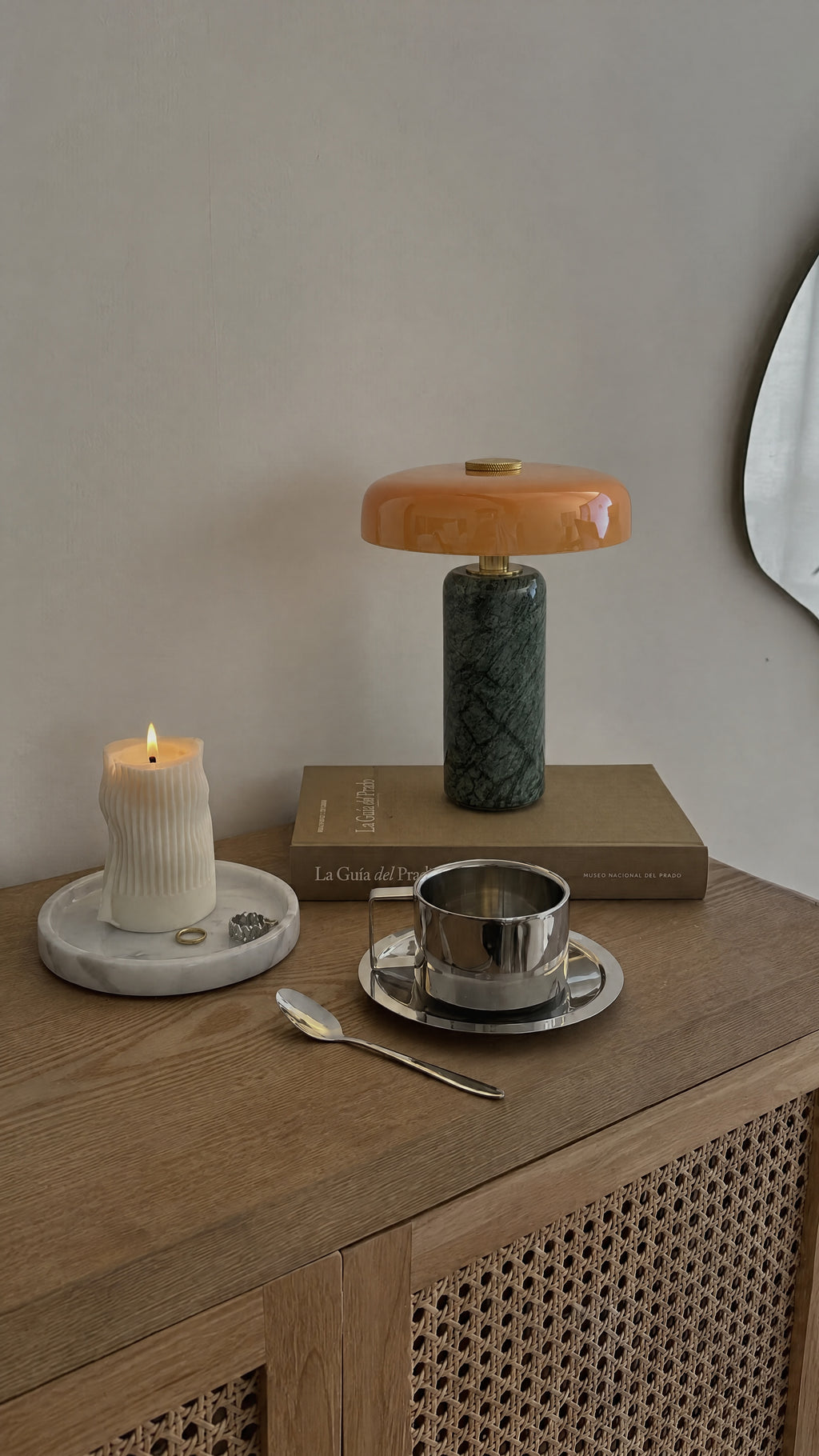 The Columba lamp unlit on a book on an oak and rattan sideboard, beside a lit pillar candle and a steel espresso cup.