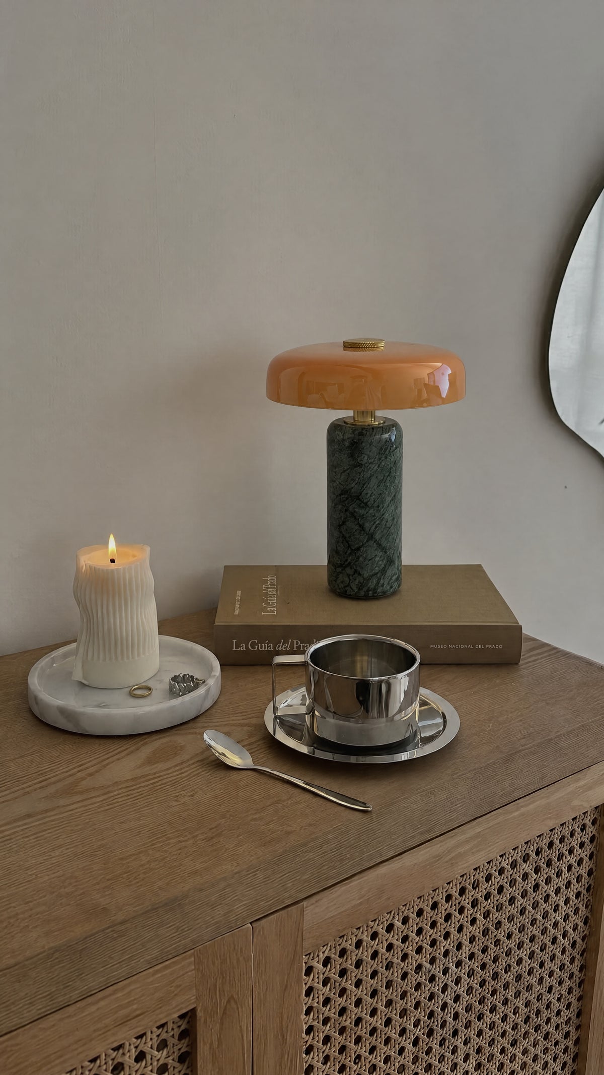 The Columba lamp unlit on a book on an oak and rattan sideboard, beside a lit pillar candle and a steel espresso cup.