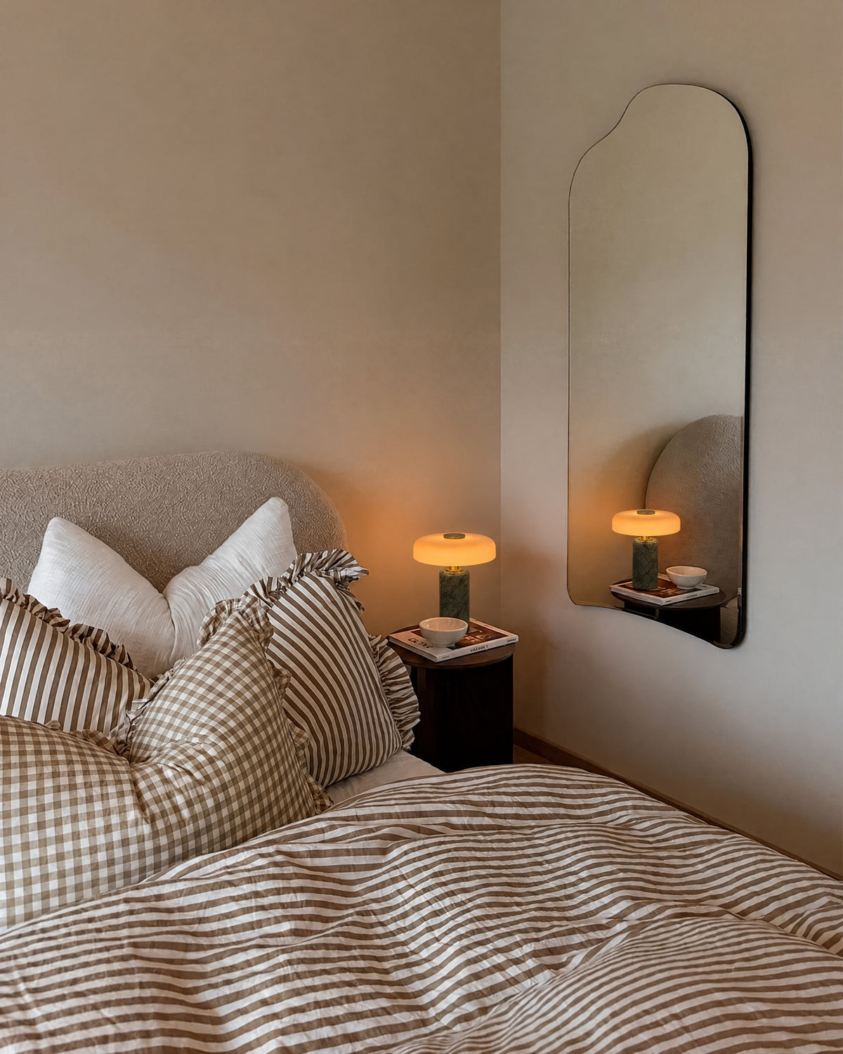 The Columba lamp lit on a bedside table next to a boucle bed with striped linen, reflected in a tall wall mirror.