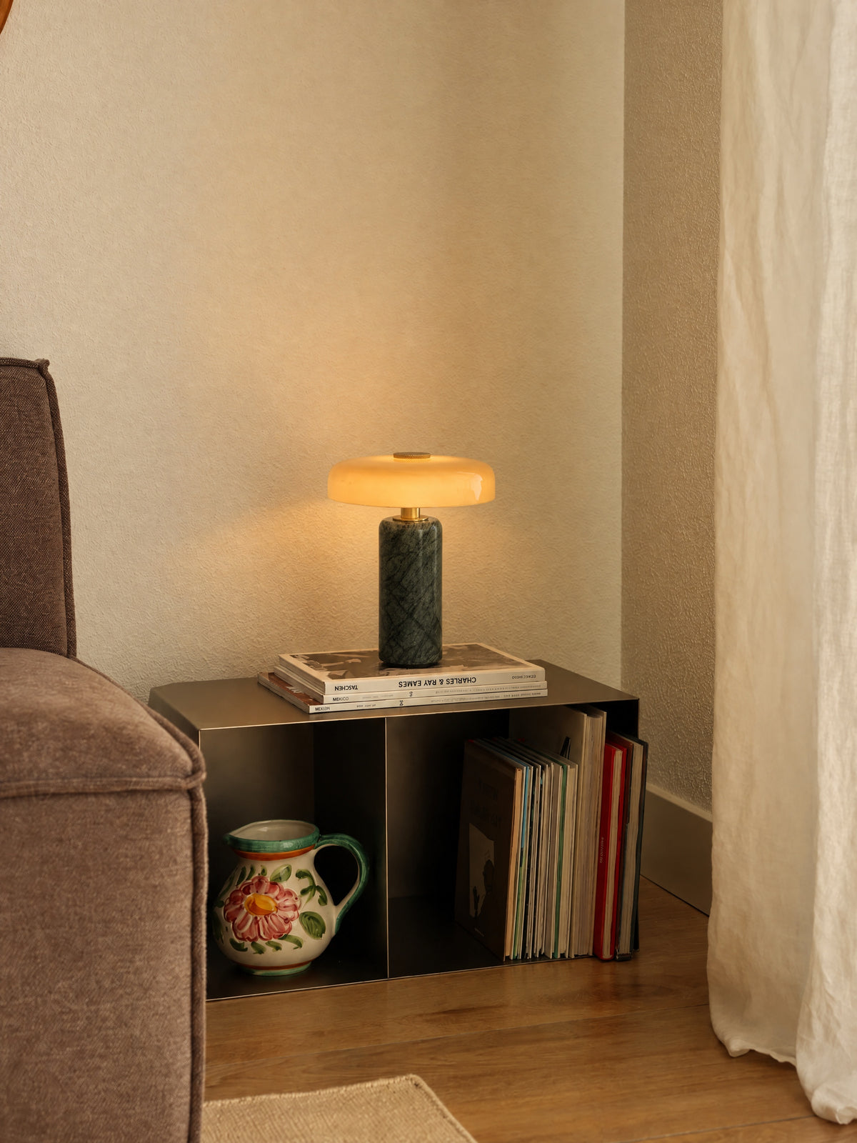 The Columba lamp lit on a stack of design books on a steel side table in a living-room corner, beside a sofa and a linen curtain.