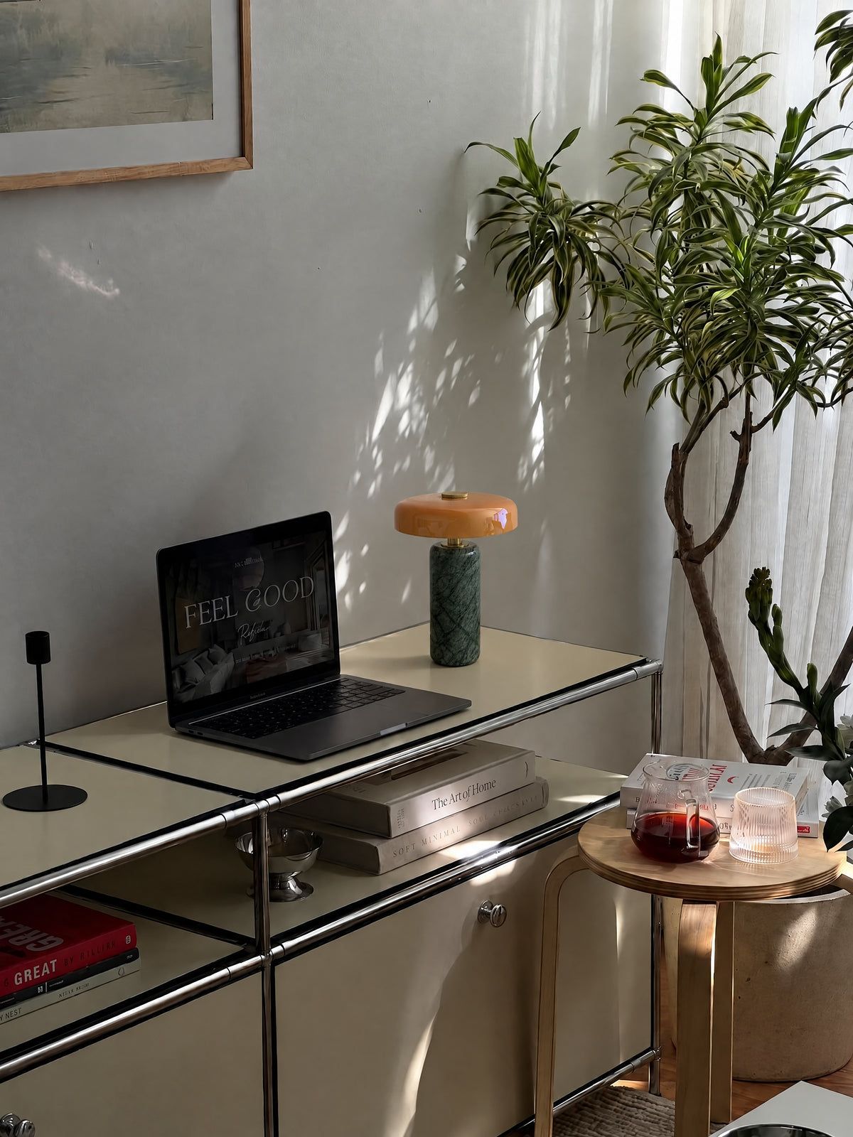 The Columba lamp unlit on a cream steel sideboard beside a laptop in daylight, with leaf shadows on the wall.
