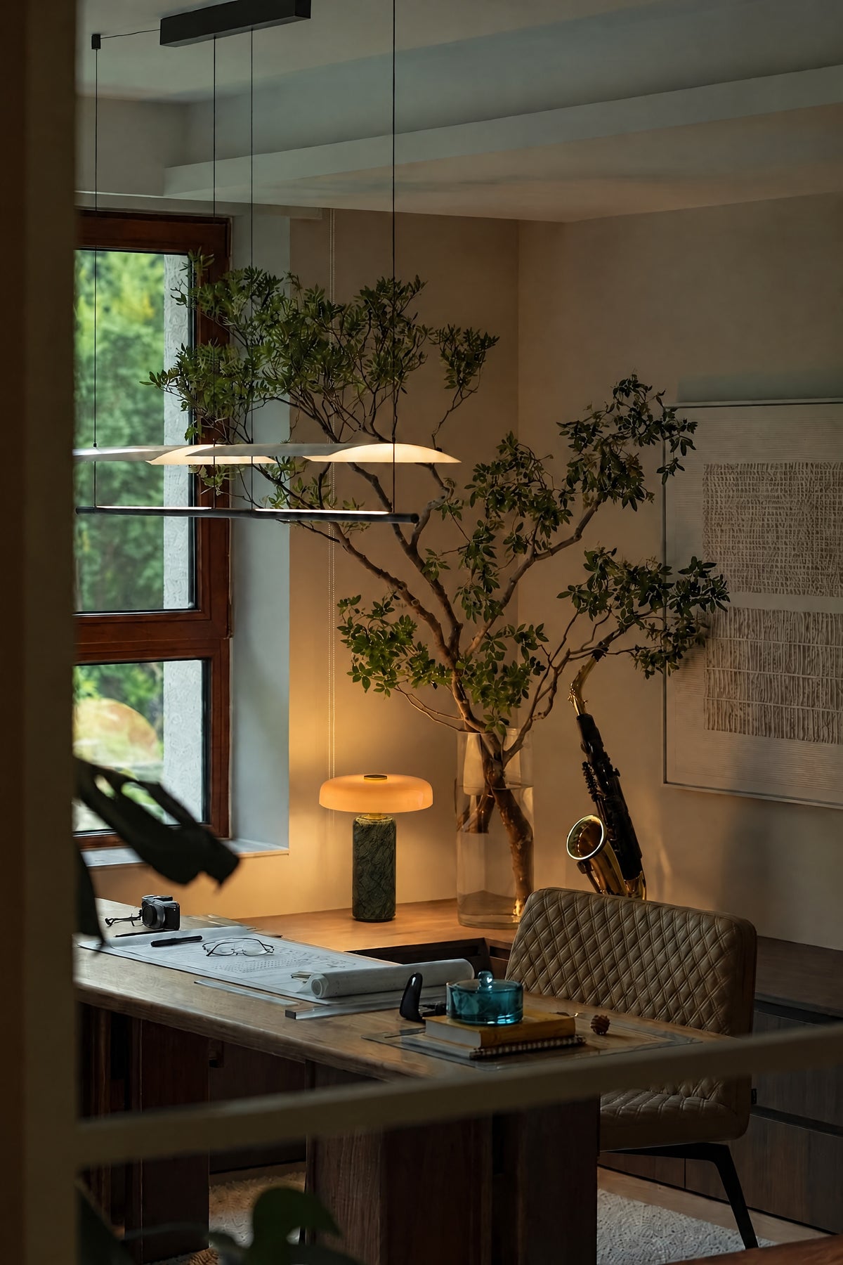 The Columba lamp lit on the desk of a quiet study at dusk, beside a tall branch in a glass vase.