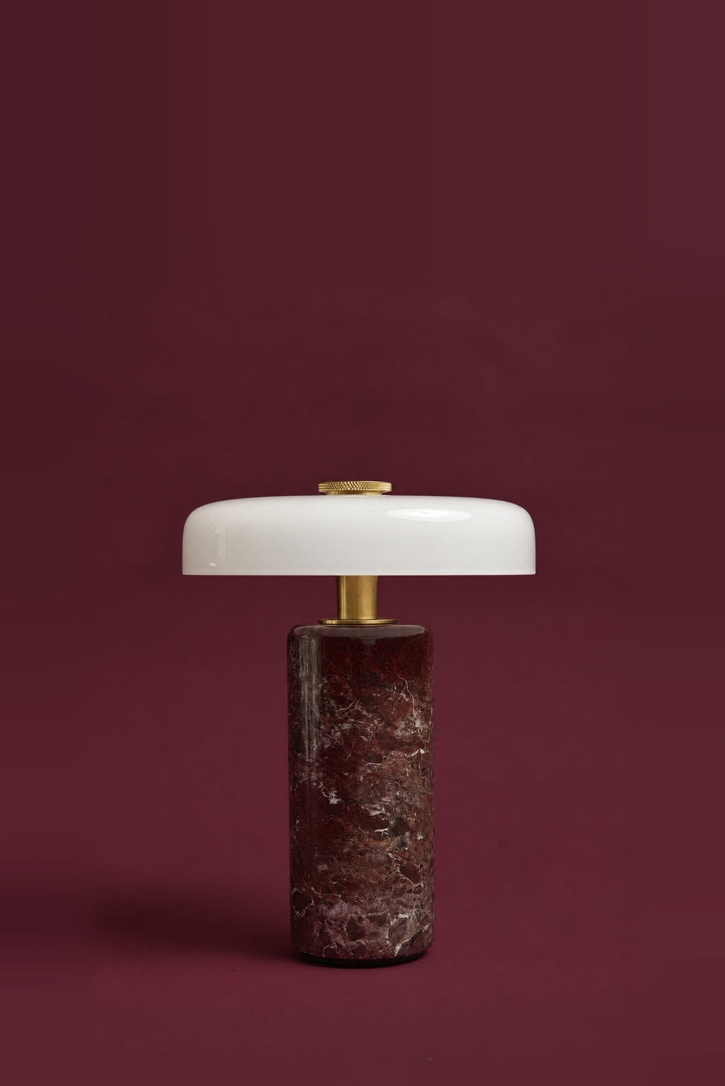 The Columba lamp, unlit: a white opal glass disc on a brass stem above a dark red Rosso Levanto marble cylinder, against a deep oxblood background.