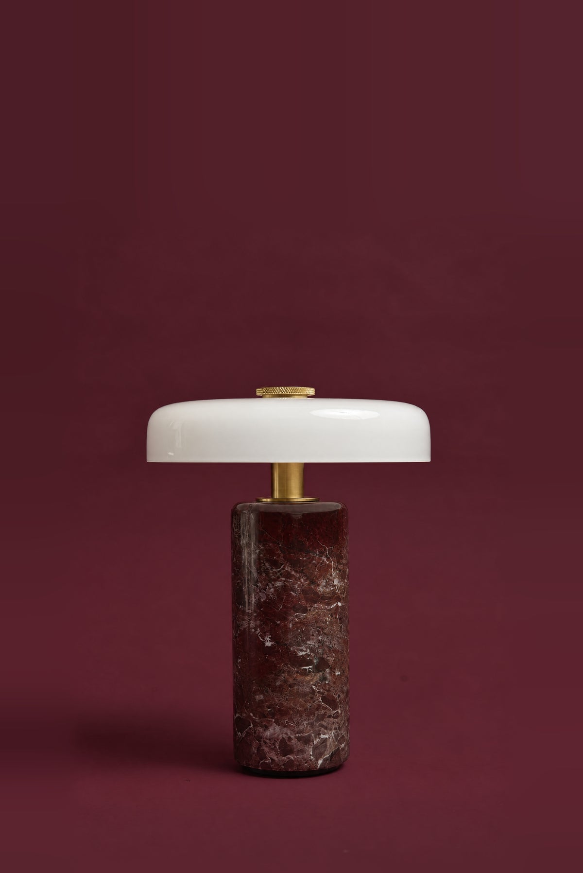 The Columba lamp, unlit: a white opal glass disc on a brass stem above a dark red Rosso Levanto marble cylinder, against a deep oxblood background.