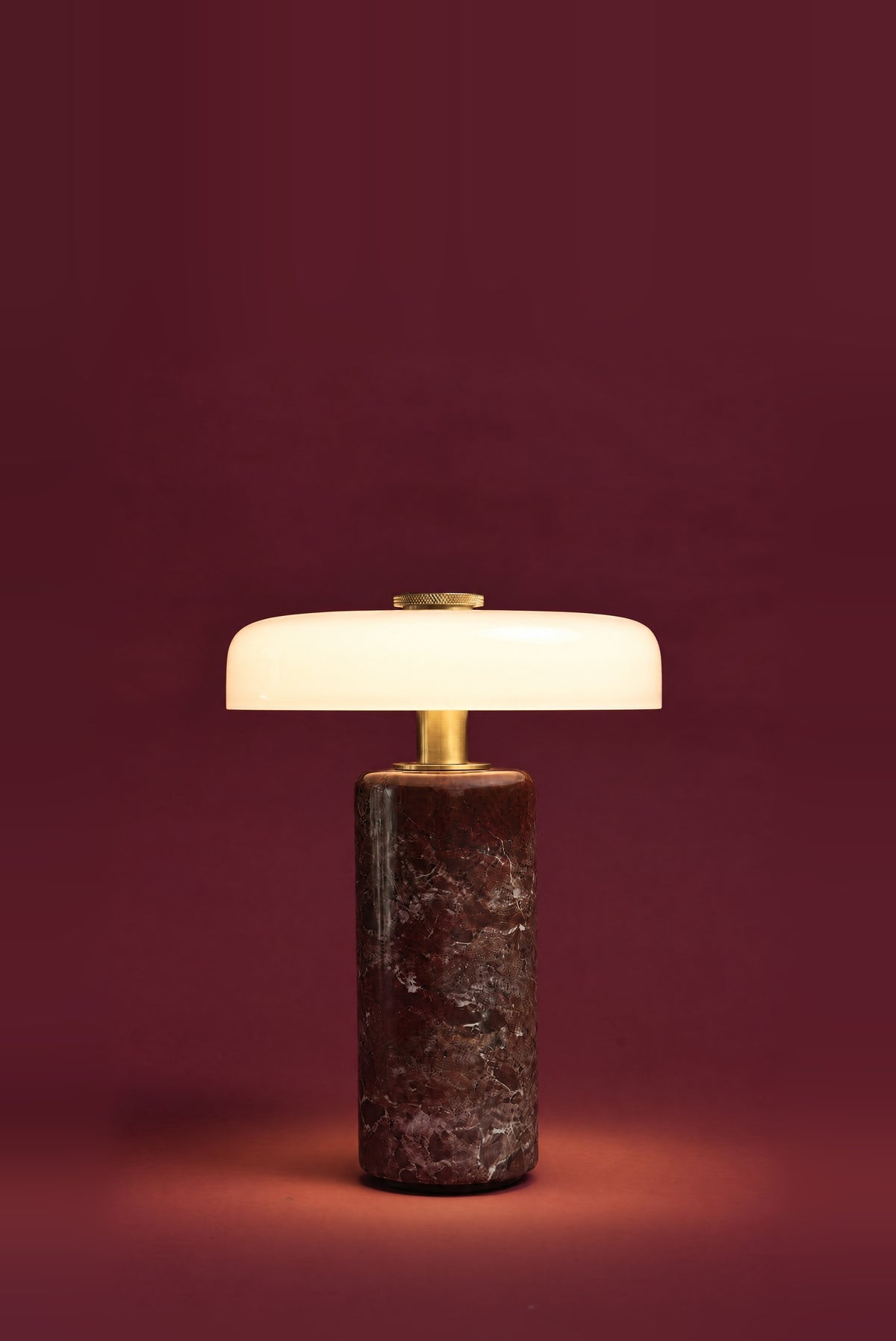 The Columba lamp, lit: the white glass disc glowing evenly, warm light pooling on the floor around the dark red marble column.