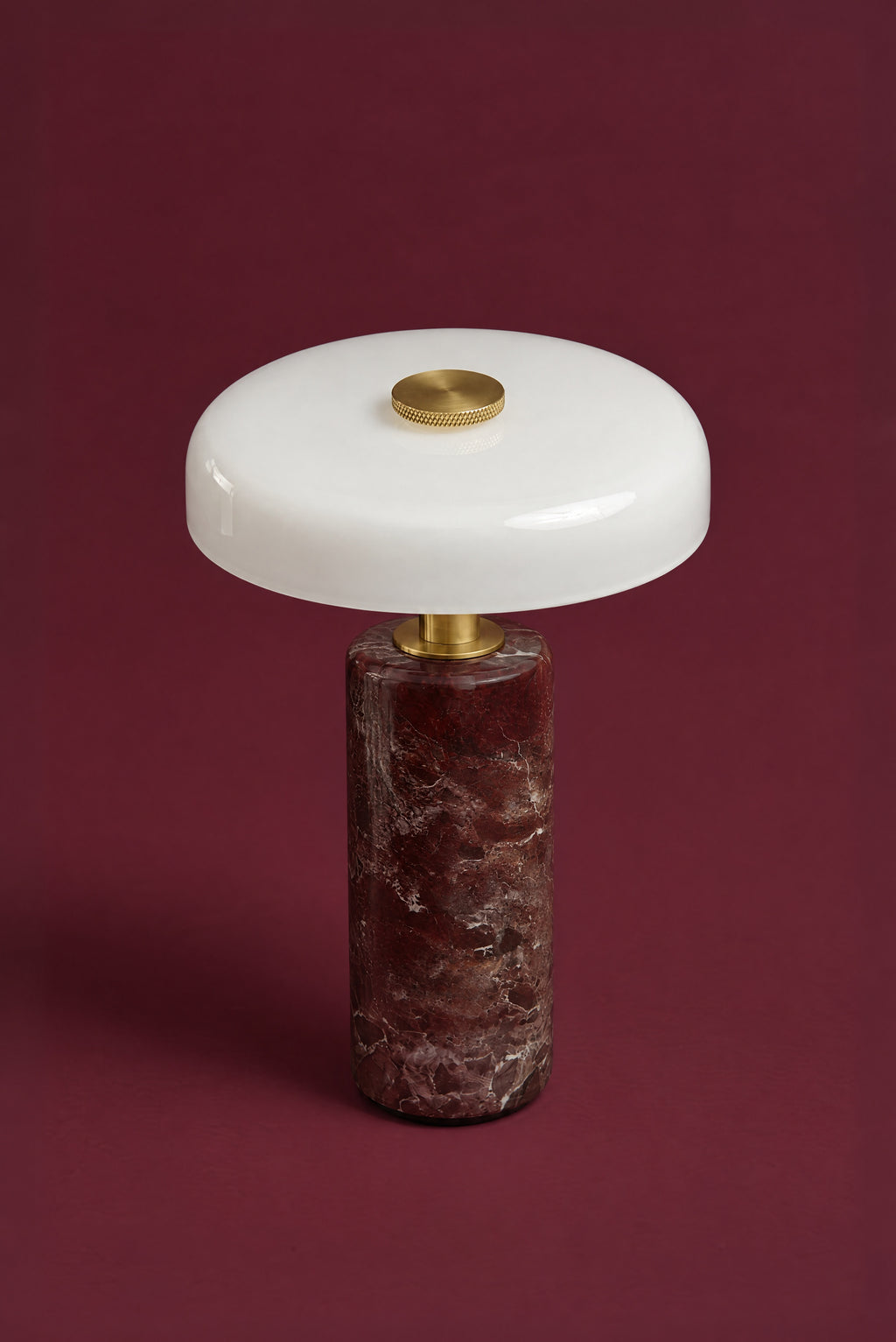 The Columba lamp in opal glass and Rosso Levanto marble from above: the glossy white disc with its knurled brass touch cap, over the dark red marble column, against a deep oxblood background.