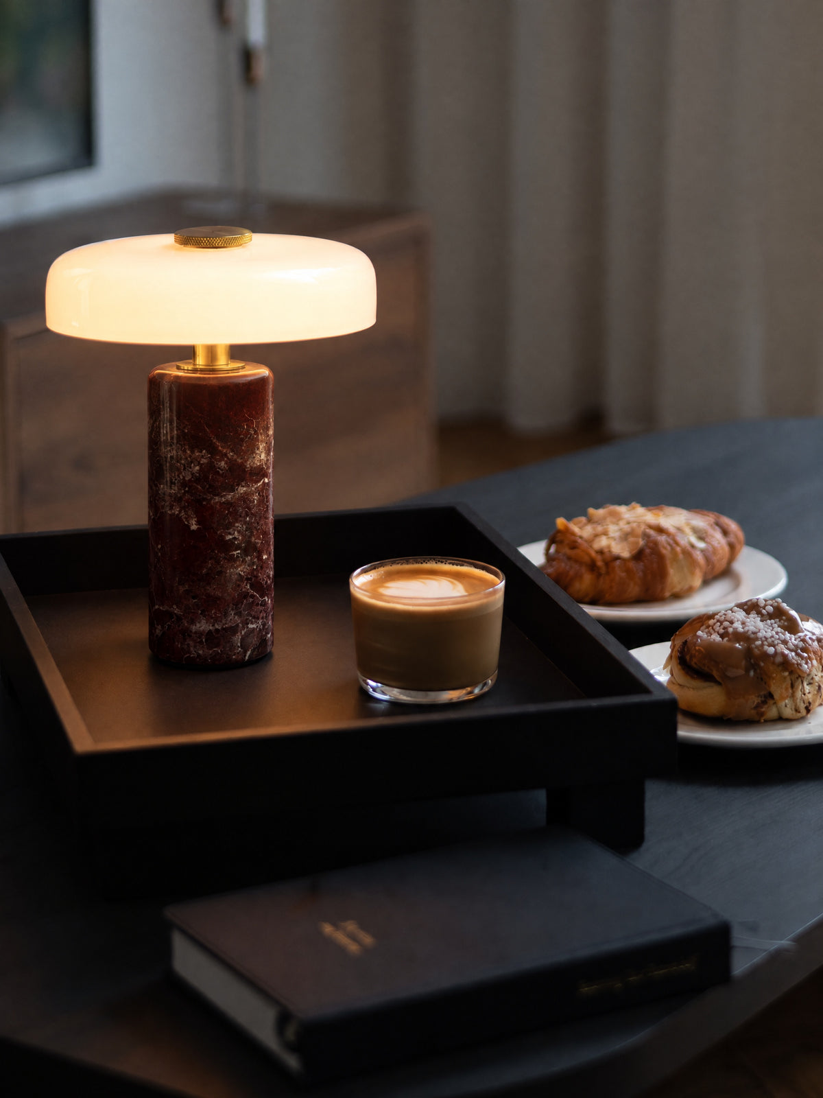 The Columba lamp lit on a black wooden tray beside a glass of flat white and pastries, warm light on the table in a dim room.