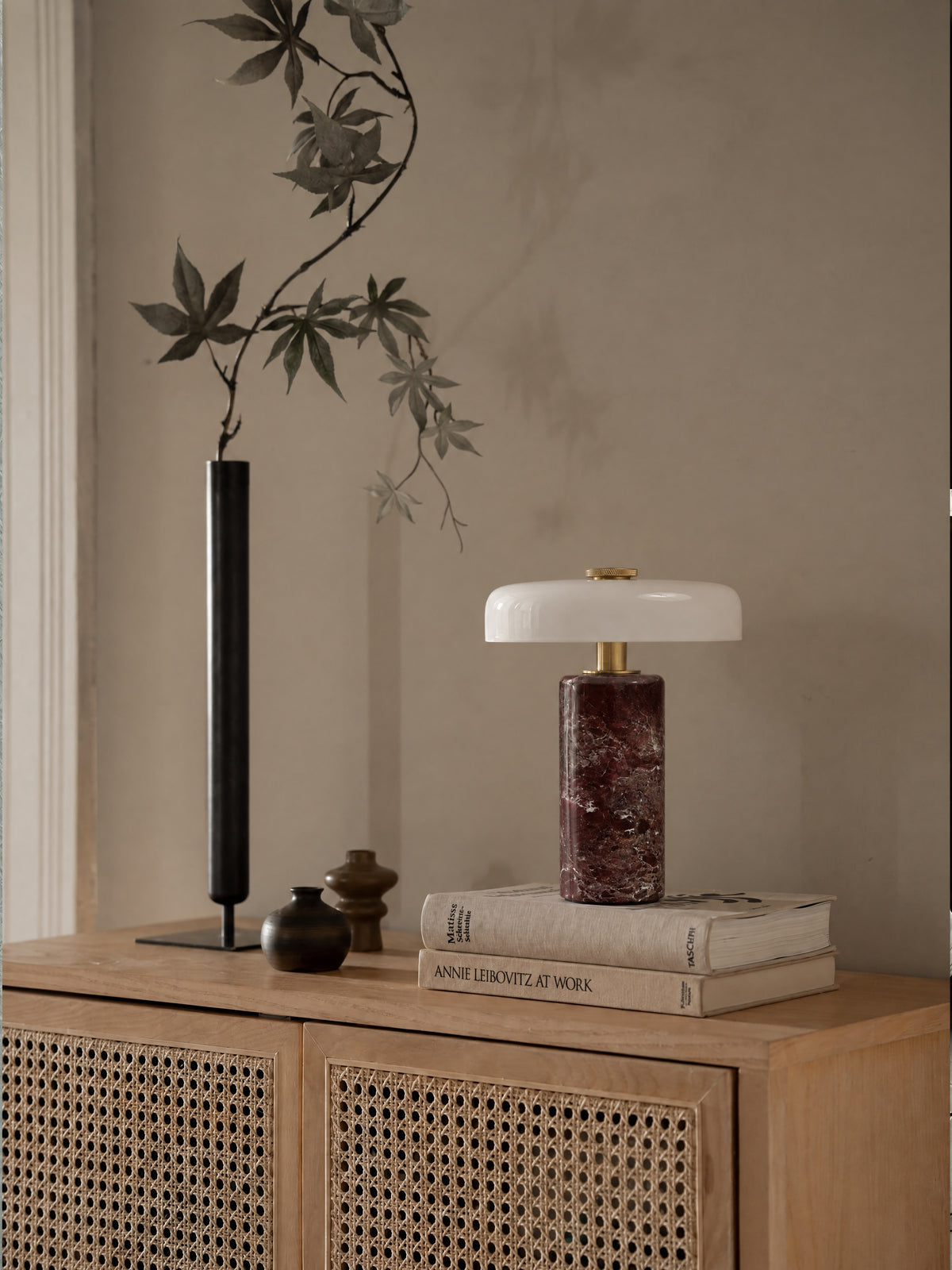 The Columba lamp unlit on a stack of books on an oak and rattan sideboard, beside a tall black bud vase with a maple branch.