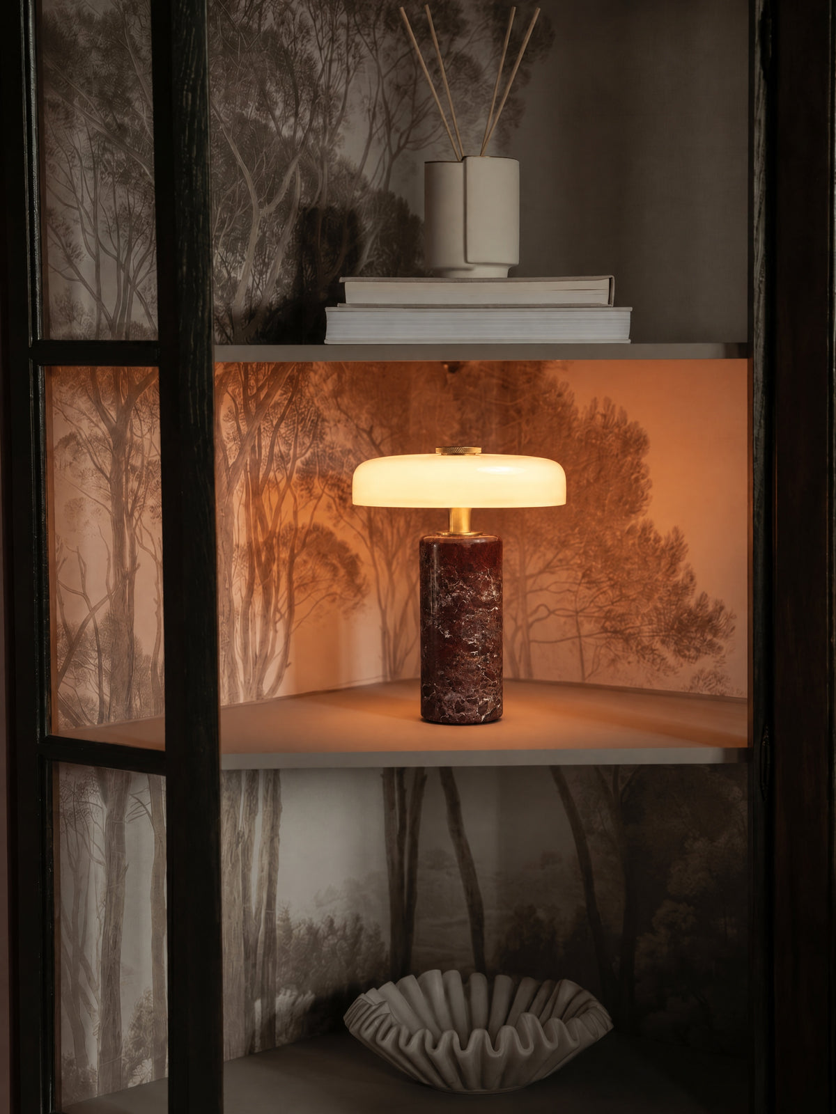 The Columba lamp lit inside a dark glass-fronted cabinet lined with forest-mural wallpaper, glowing warm on the middle shelf.